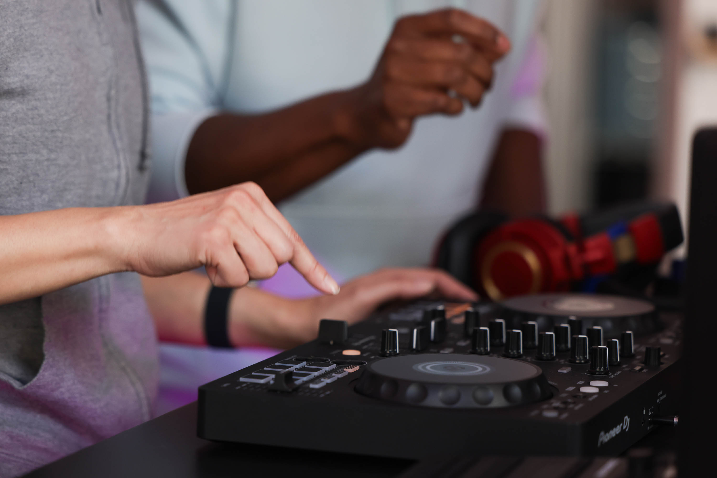 Two people operate a DJ mixing console with knobs and buttons; headphones lie in the background.