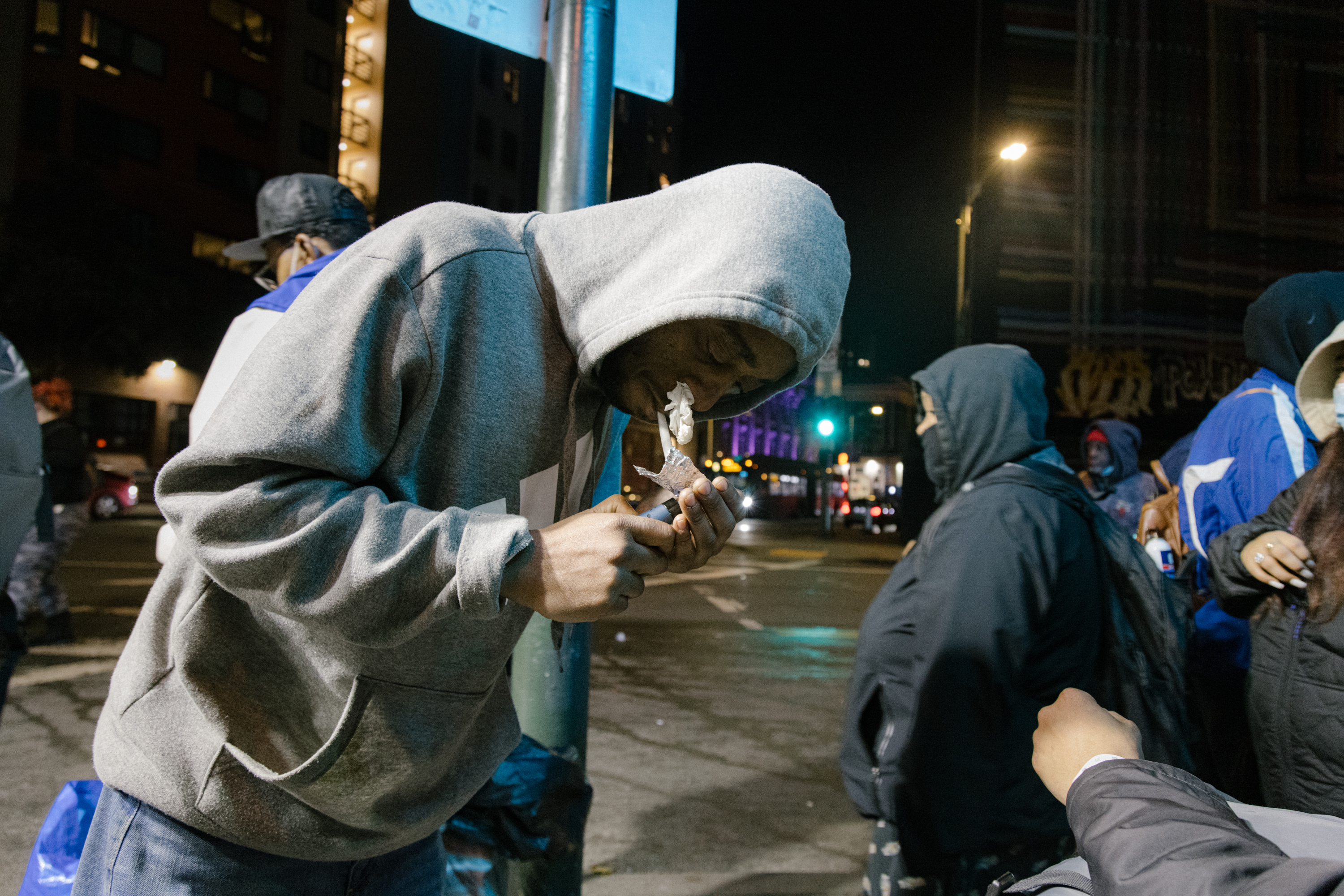 A person in a hoodie is lighting a pipe on a city street at night, surrounded by others in winter attire.