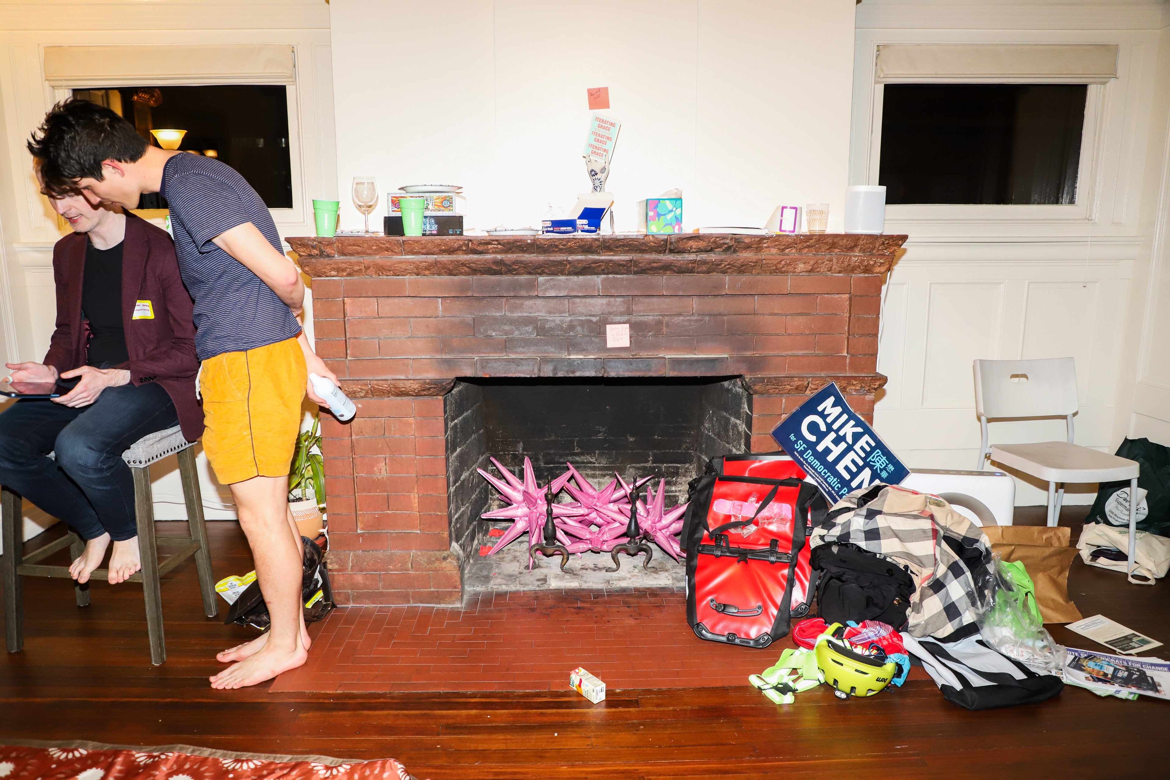 Scattered personal belongings and a Mike Chen campaign sign sit in front of a brink fireplace in a cluttered room with two individuals.