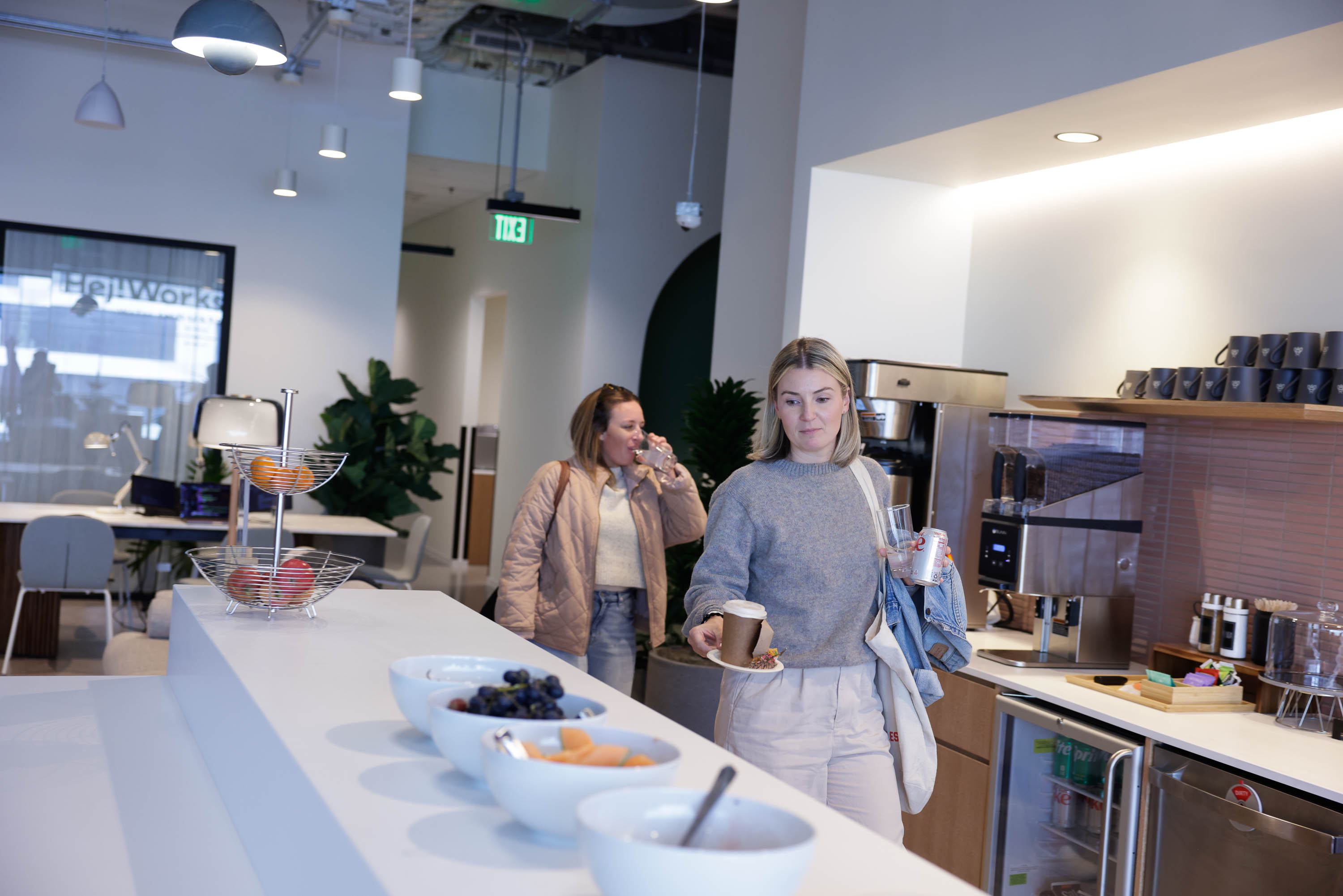 Two women at a modern office kitchenette with fruit bowls and a coffee machine.