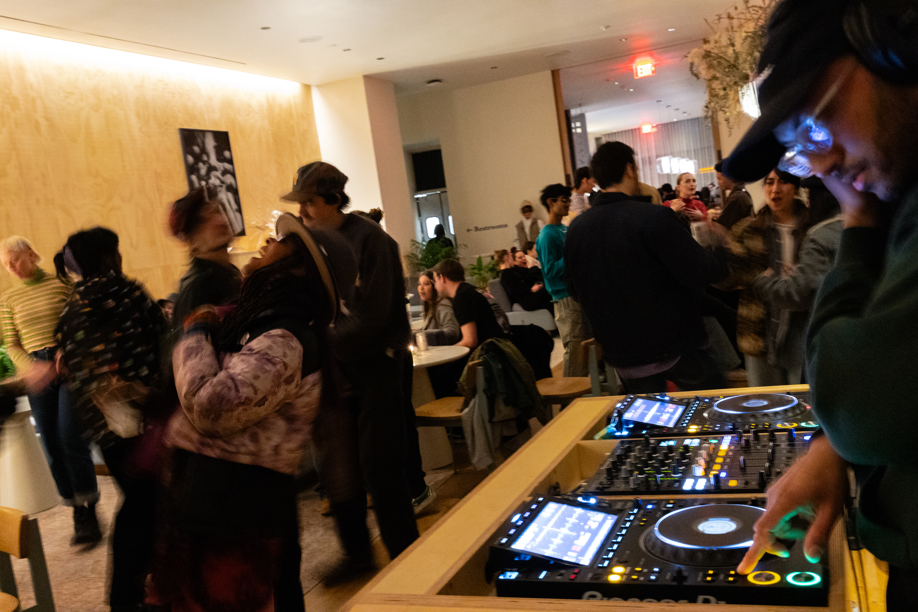 A DJ at a mixer with people mingling in a dimly lit indoor space.