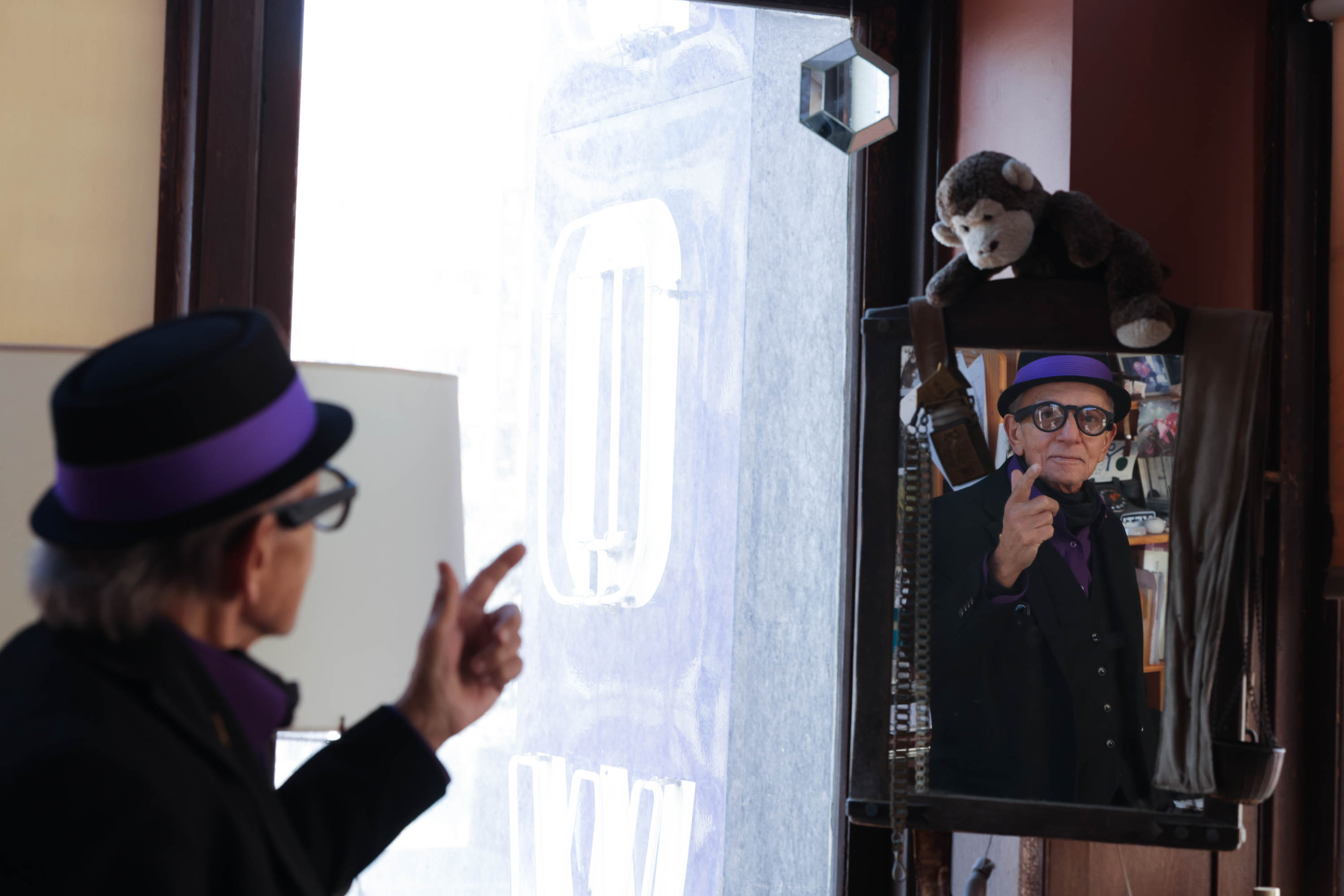A person in a purple hat gestures, reflected in a mirror beside a plush monkey. Bright light shines from a window.