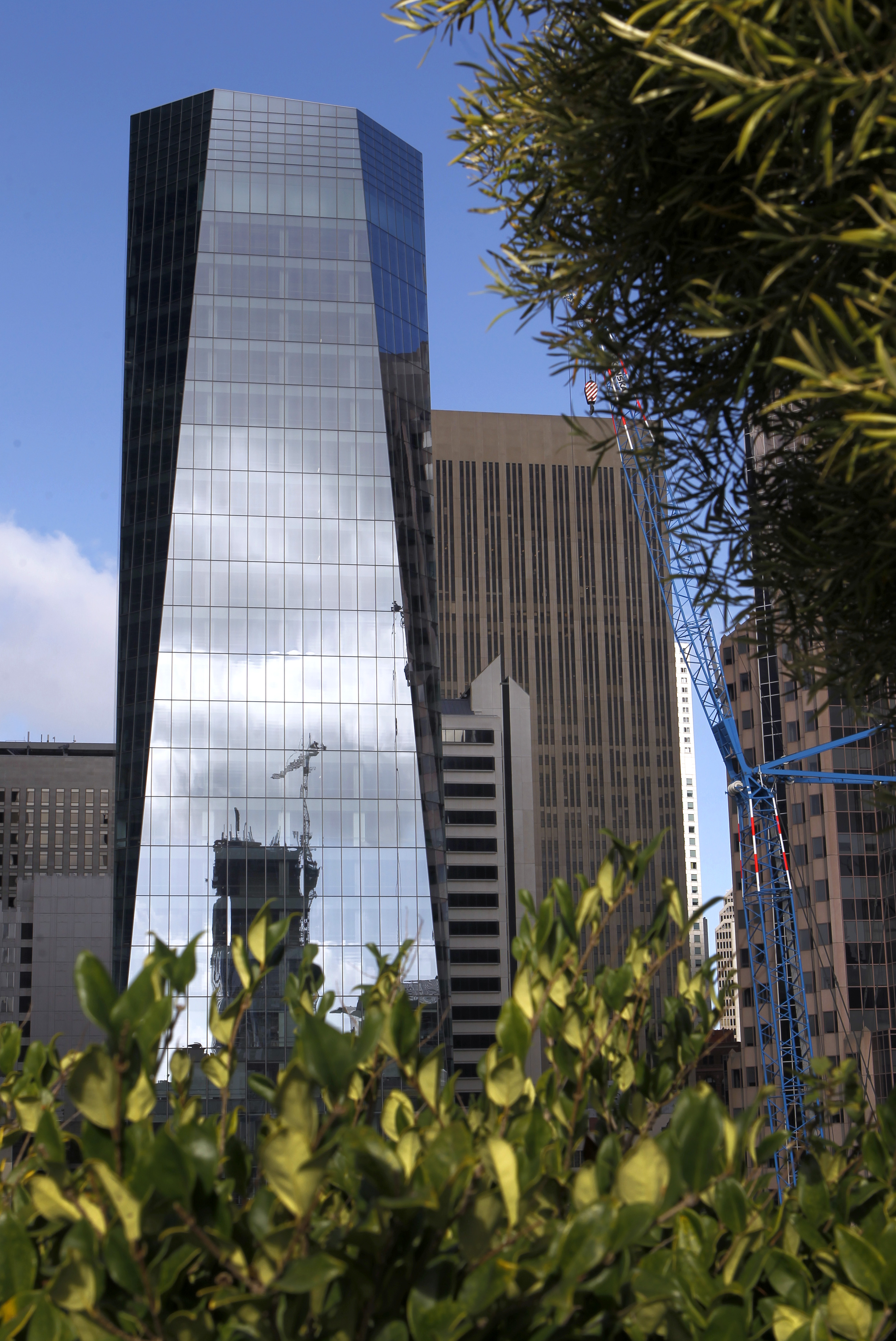 A reflective skyscraper towers over other buildings, with foliage in the foreground and a crane visible in the reflection.