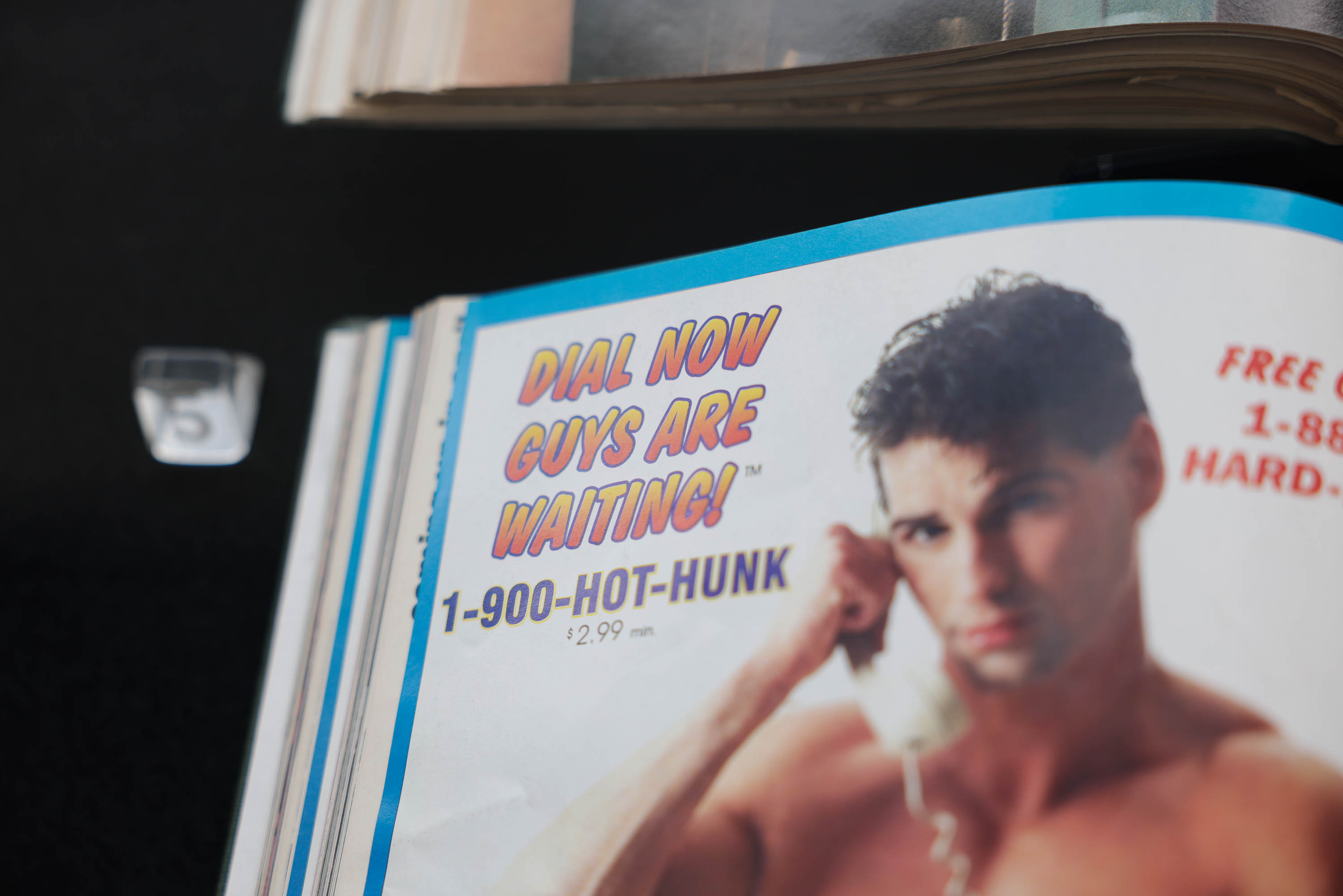 An ad with a shirtless man holding a phone reads &quot;DIAL NOW GUYS ARE WAITING! 1-900-HOT-HUNK $2.99/min.&quot;