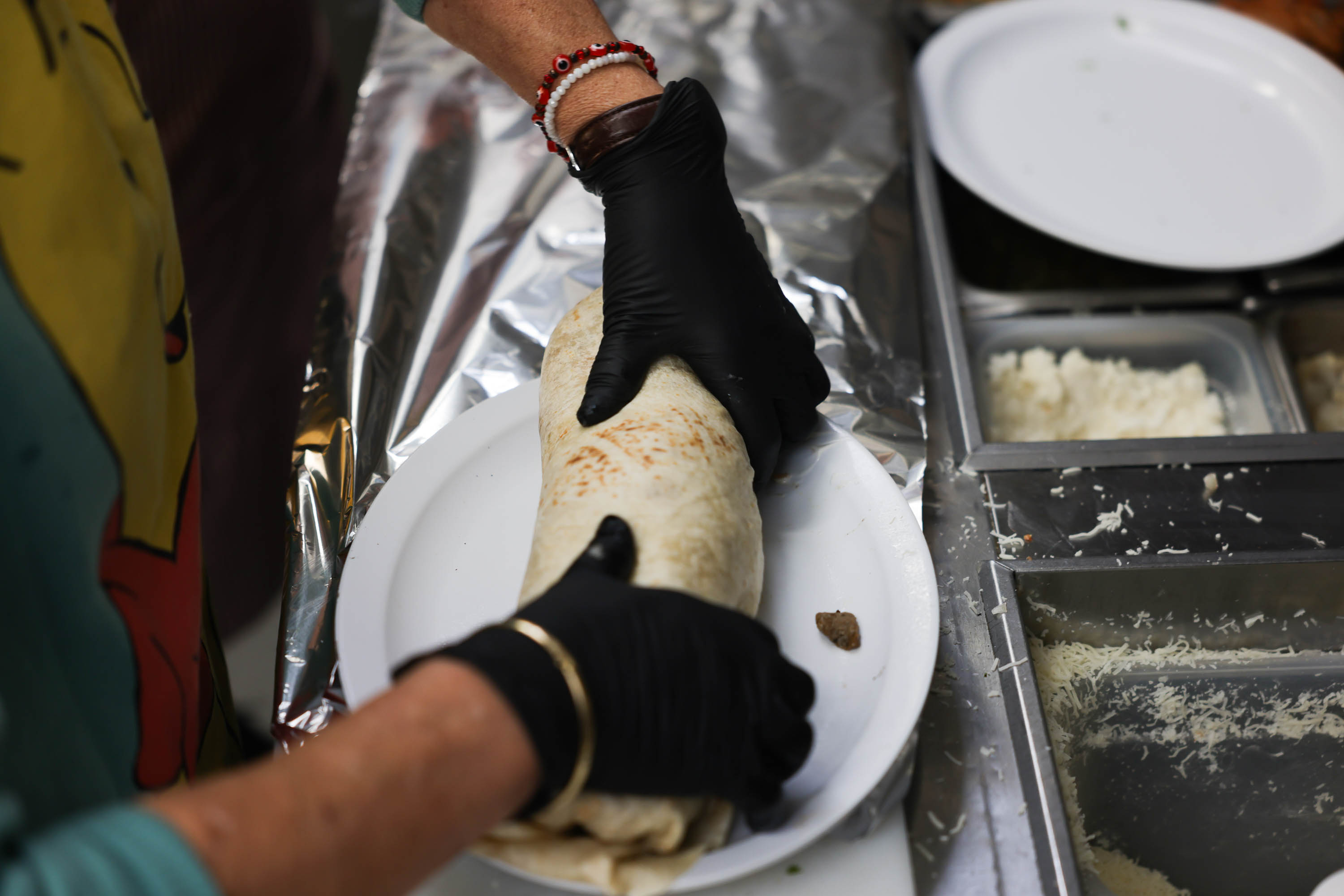 Hands in black gloves roll a burrito on a white plate in a kitchen with ingredients nearby.