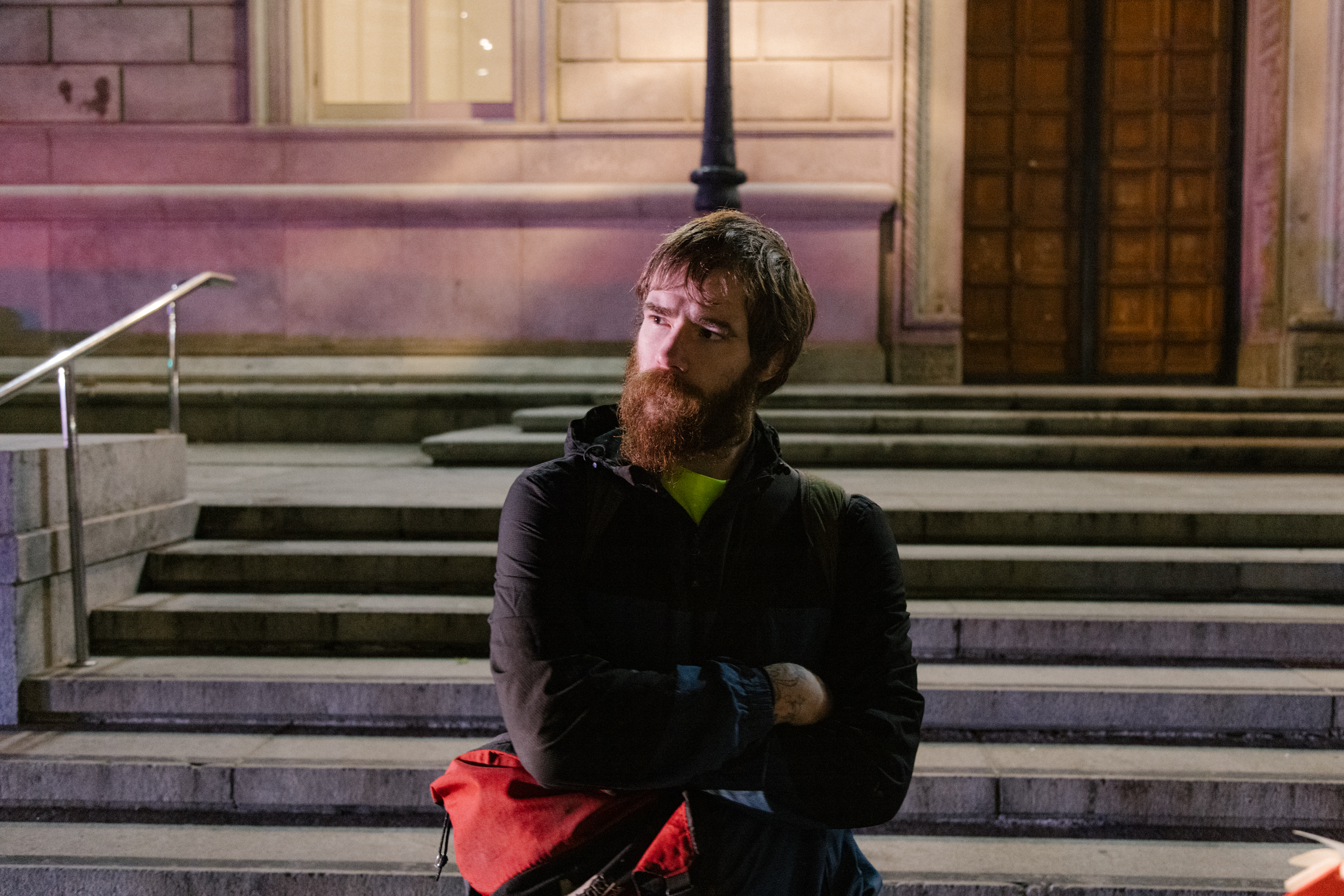 A bearded man carrying a red bag stands before a building at night, arms crossed, deep in thought.