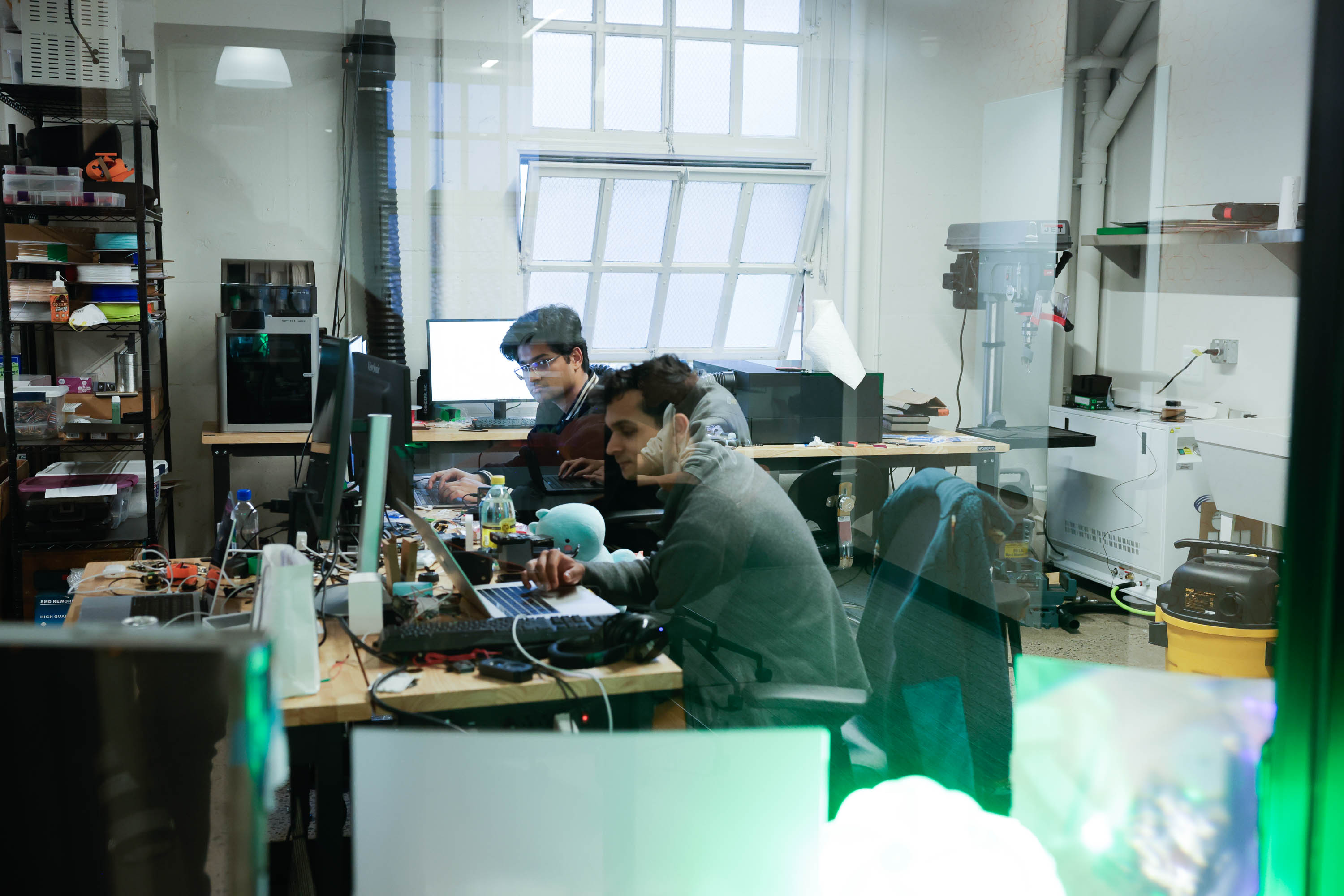 Two people work at cluttered desks with electronic equipment in a workshop-like room.