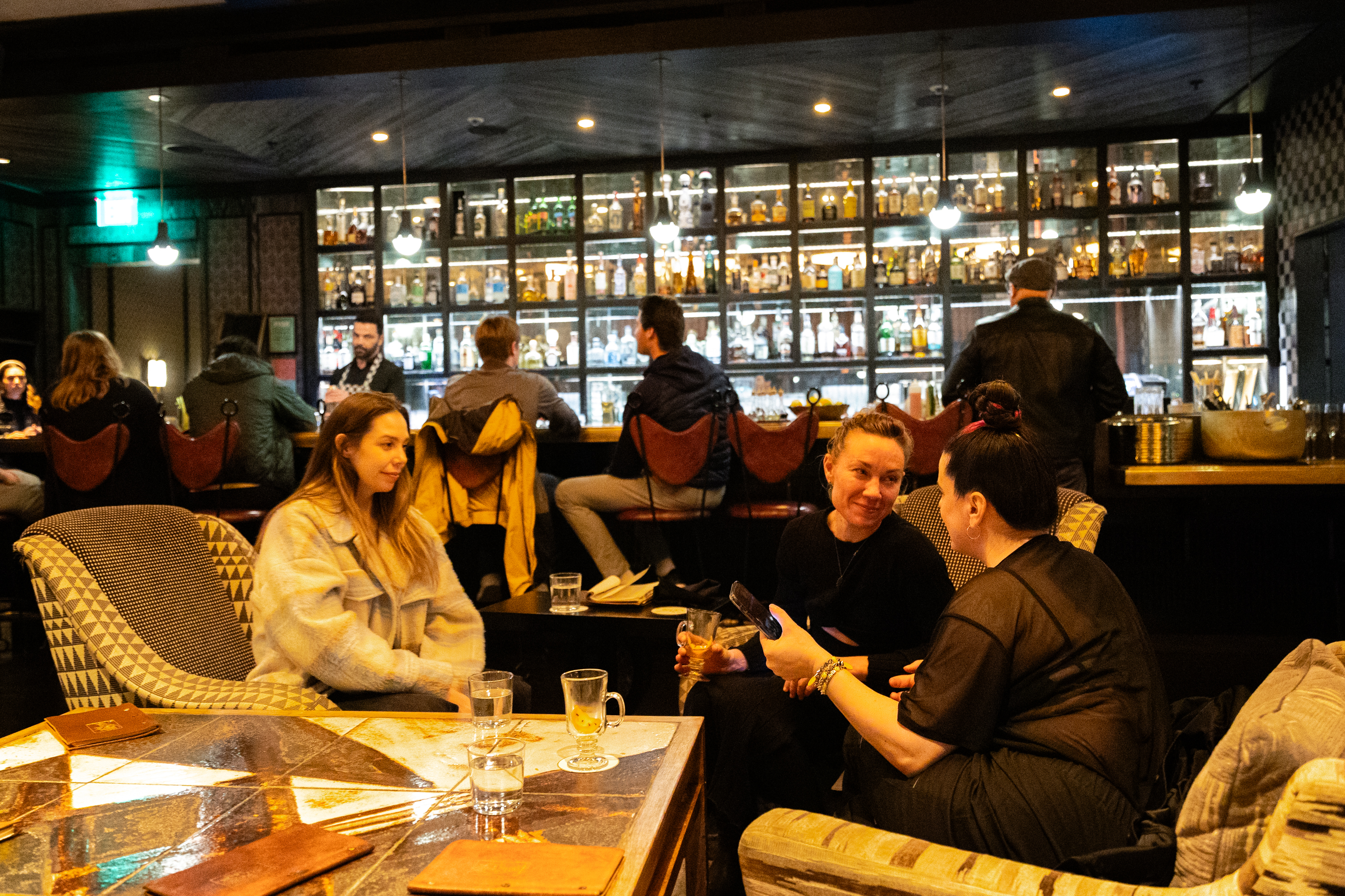 People socializing in a cozy bar with a backlit shelf of bottles.