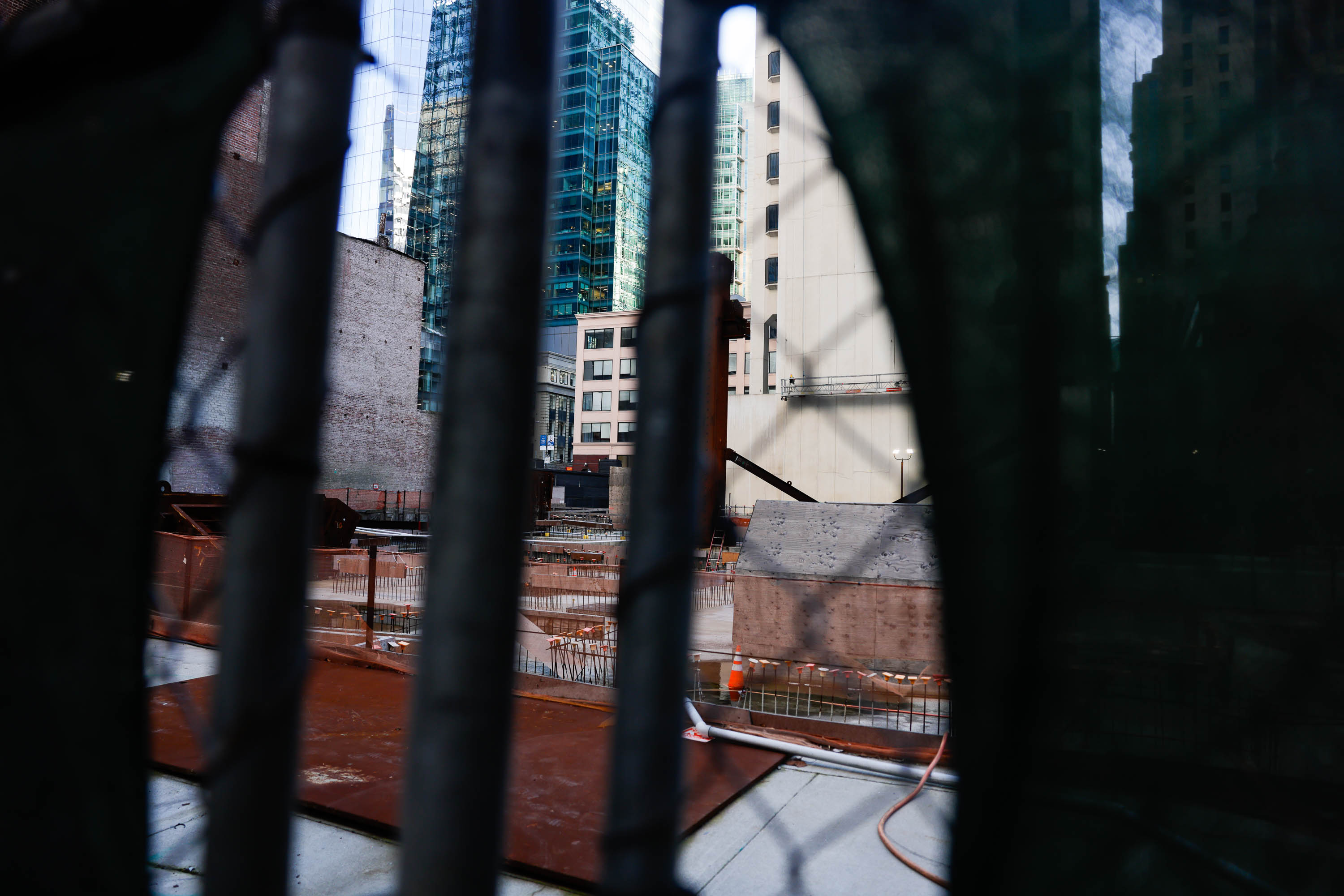 A construction site viewed through a fence, with urban buildings and skyscrapers in the background.