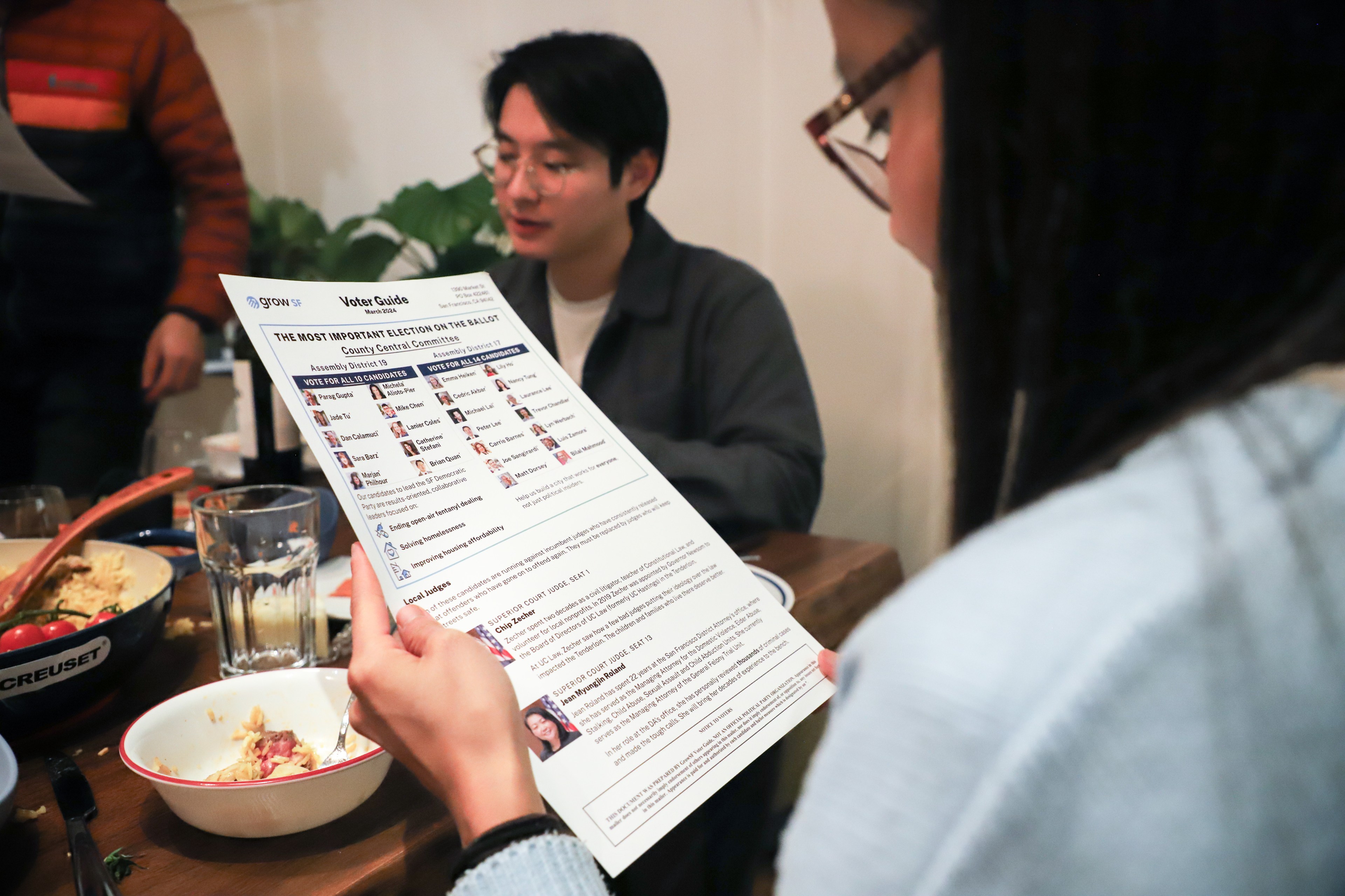 A person reads a voter guide at a table where others are engaged in discussion, with food and drinks present.
