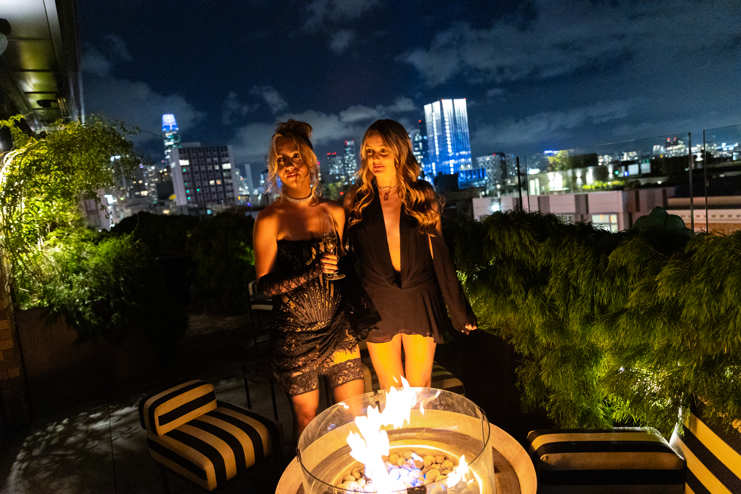 Two people stand by a fire pit on a rooftop at night with city lights in the background.