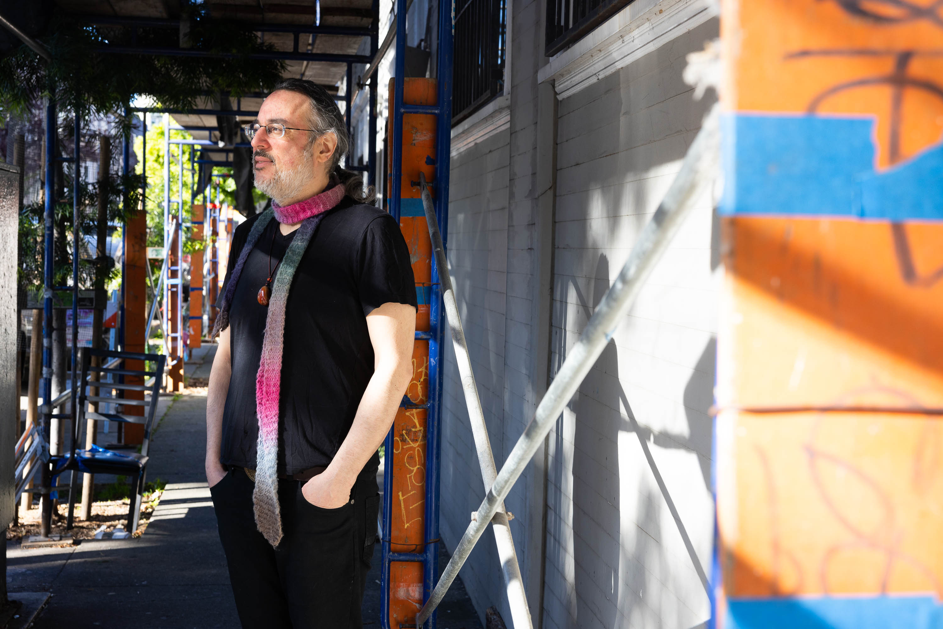 A man stands by a scaffolding in a sunlit alley, wearing a black shirt and pink scarf.
