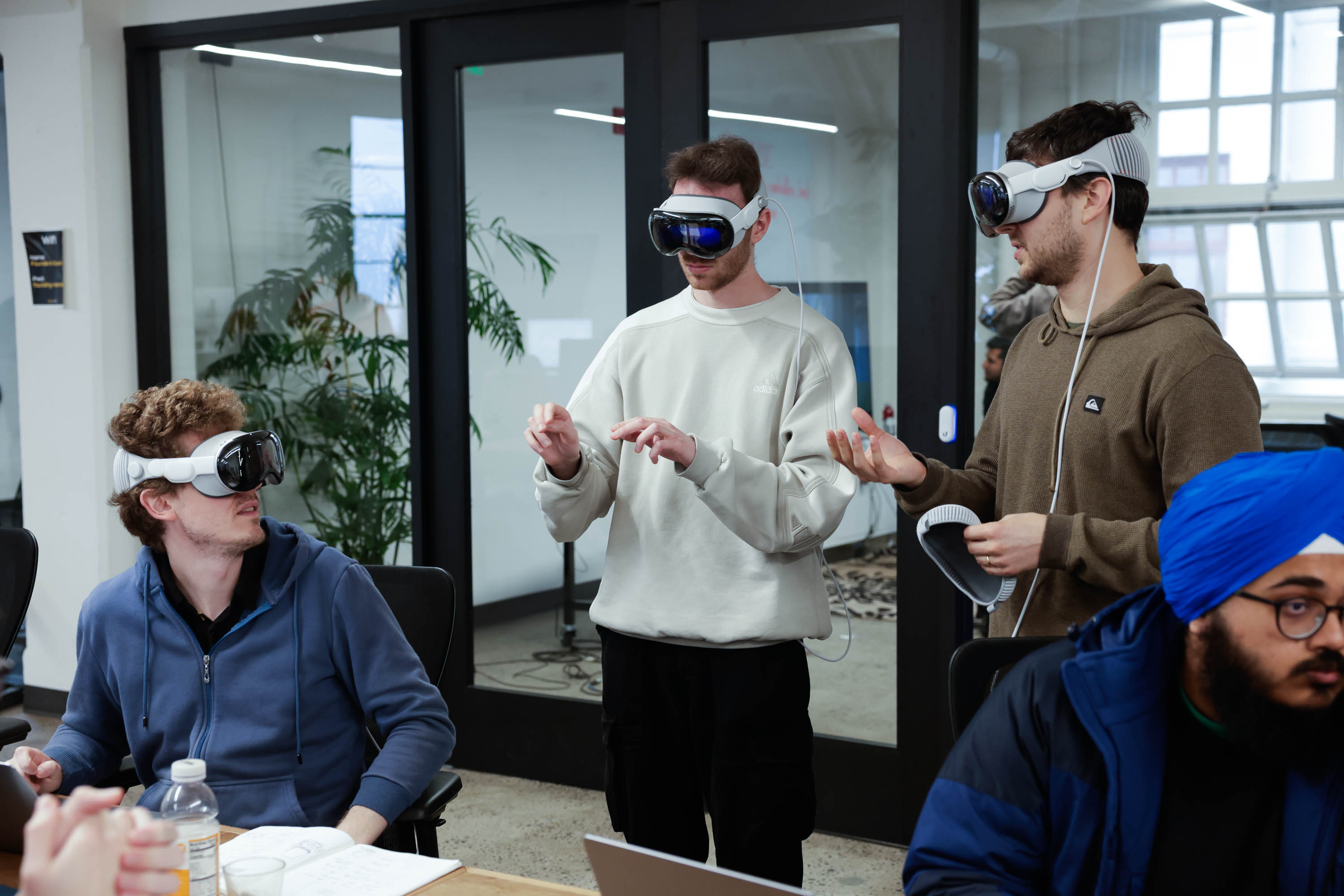 Three men wear Apple Vision Pro headsets and gesture in an office-like setting with a seated colleague nearby.