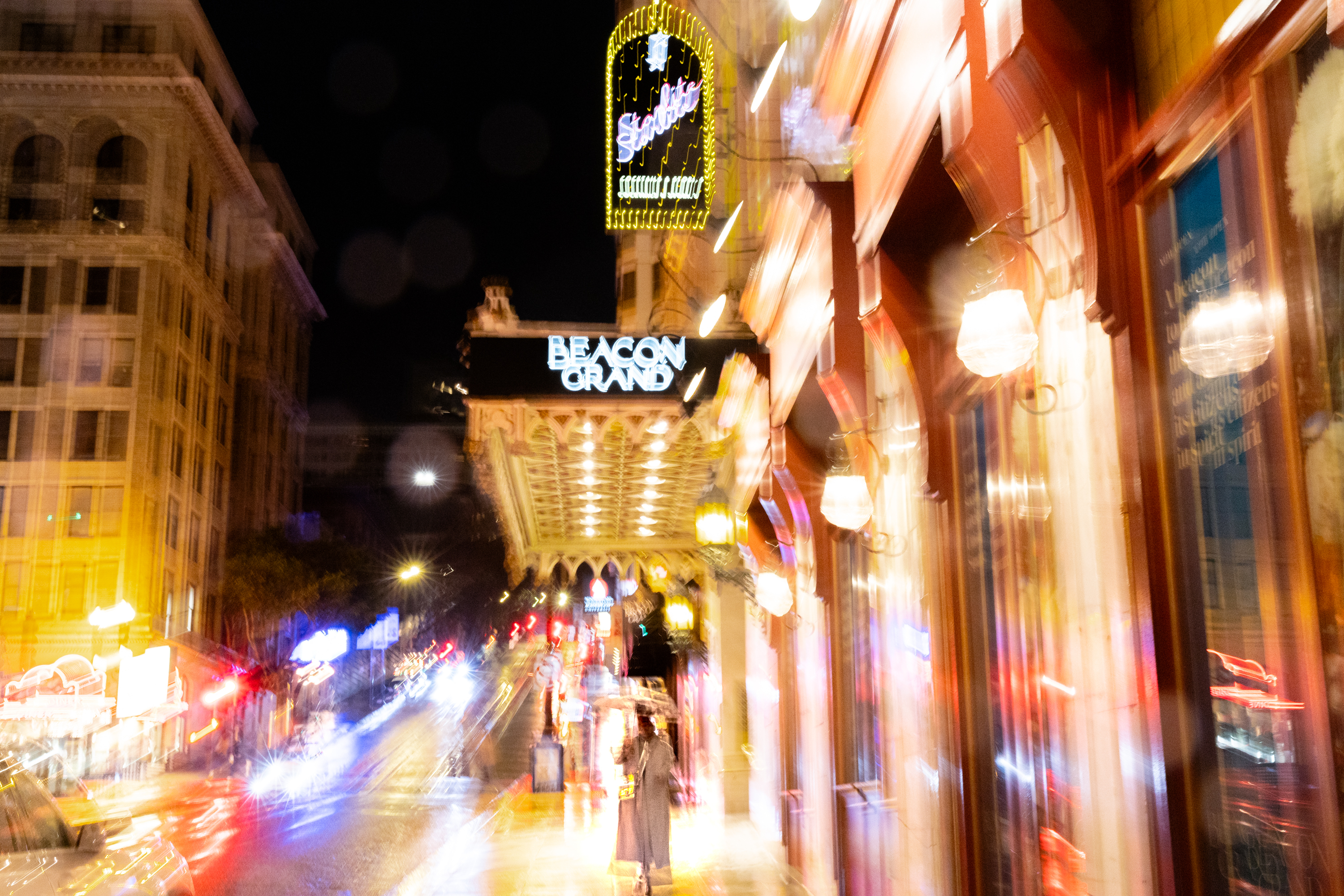 A blurry night scene with bright lights, a neon sign, and &quot;BEACON GRAND&quot; sign above an illuminated entrance.