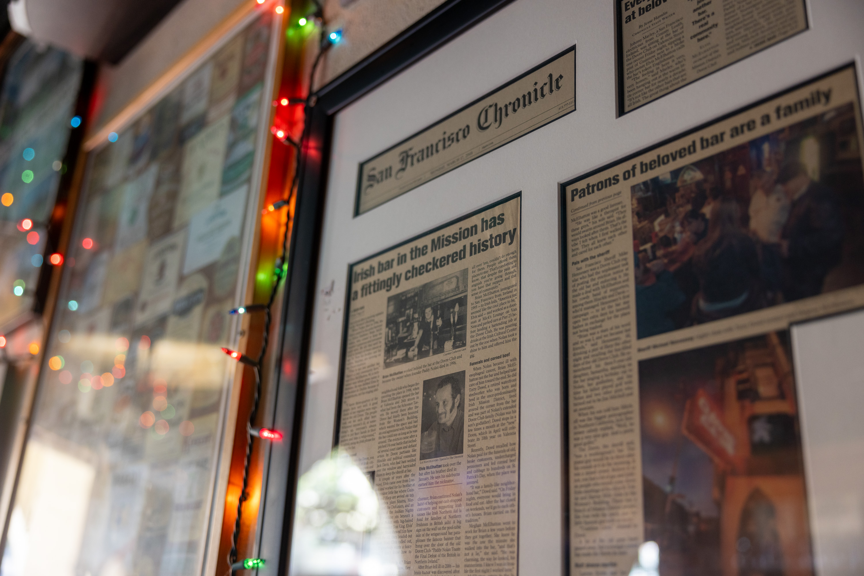 Newspaper clippings about an Irish bar are framed on a wall, adorned with small multicolored lights.