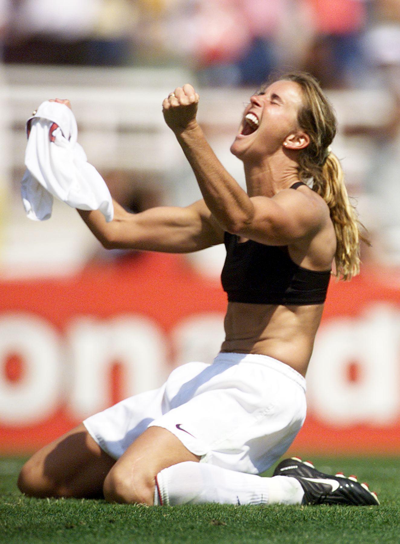 A joyful athlete on her knees on a soccer field, shouting in triumph, with a jersey in hand.