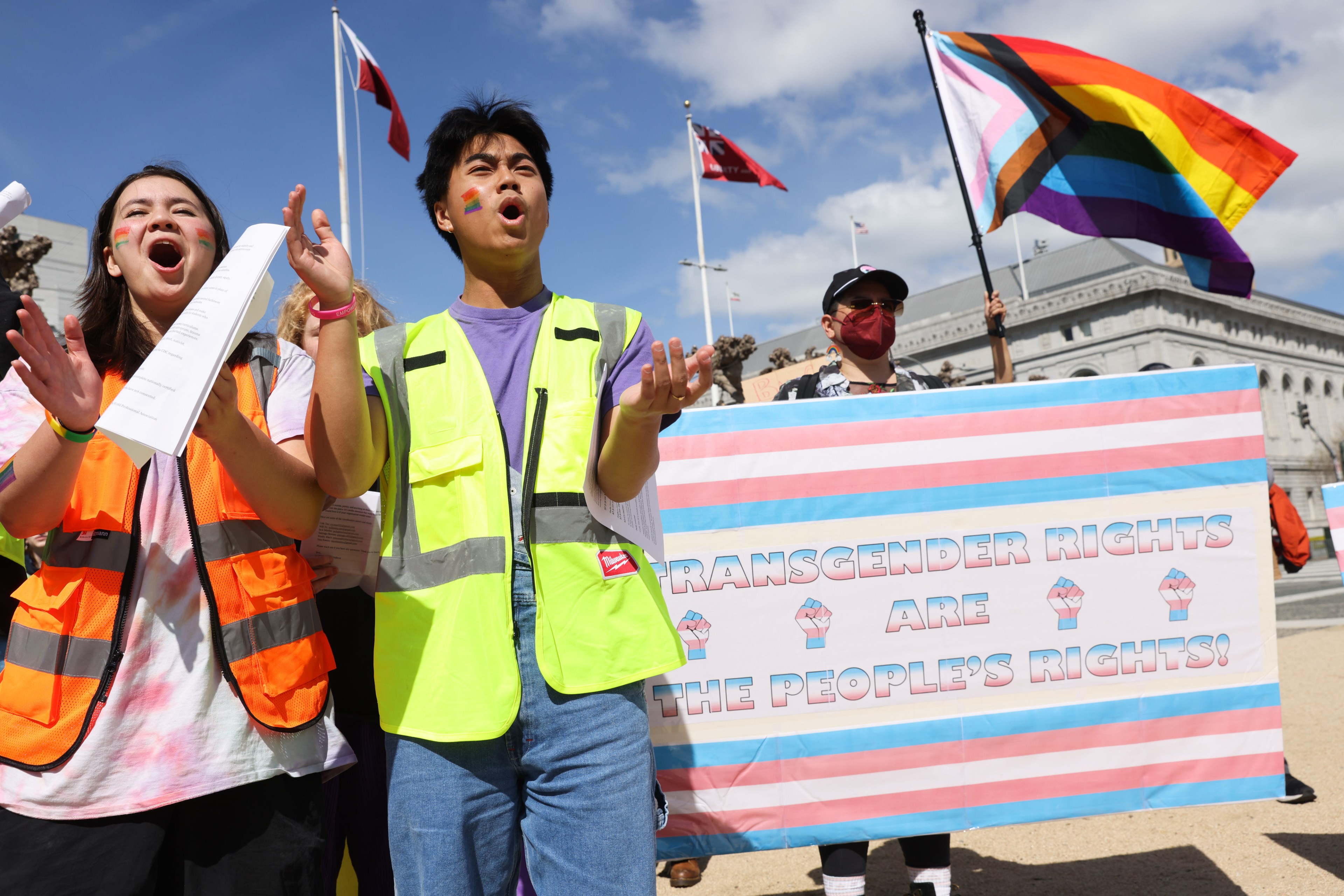 shot from below, three young people in safety vests chant and hold up a trans flag protest sign with a Progress Flag behind them.