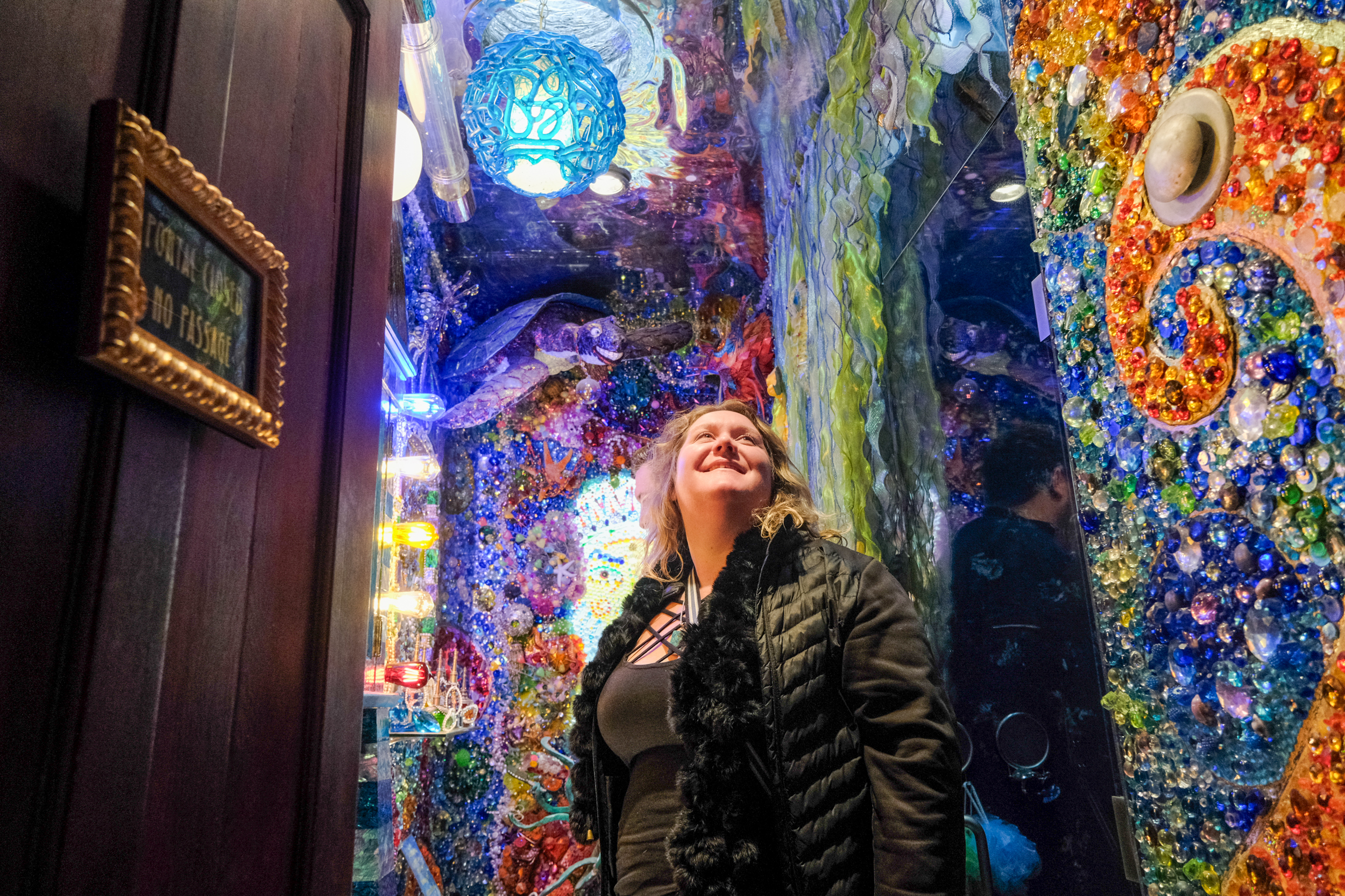 A woman looks up, smiling, in a vibrant, eclectic hallway adorned with colorful glass and lights.