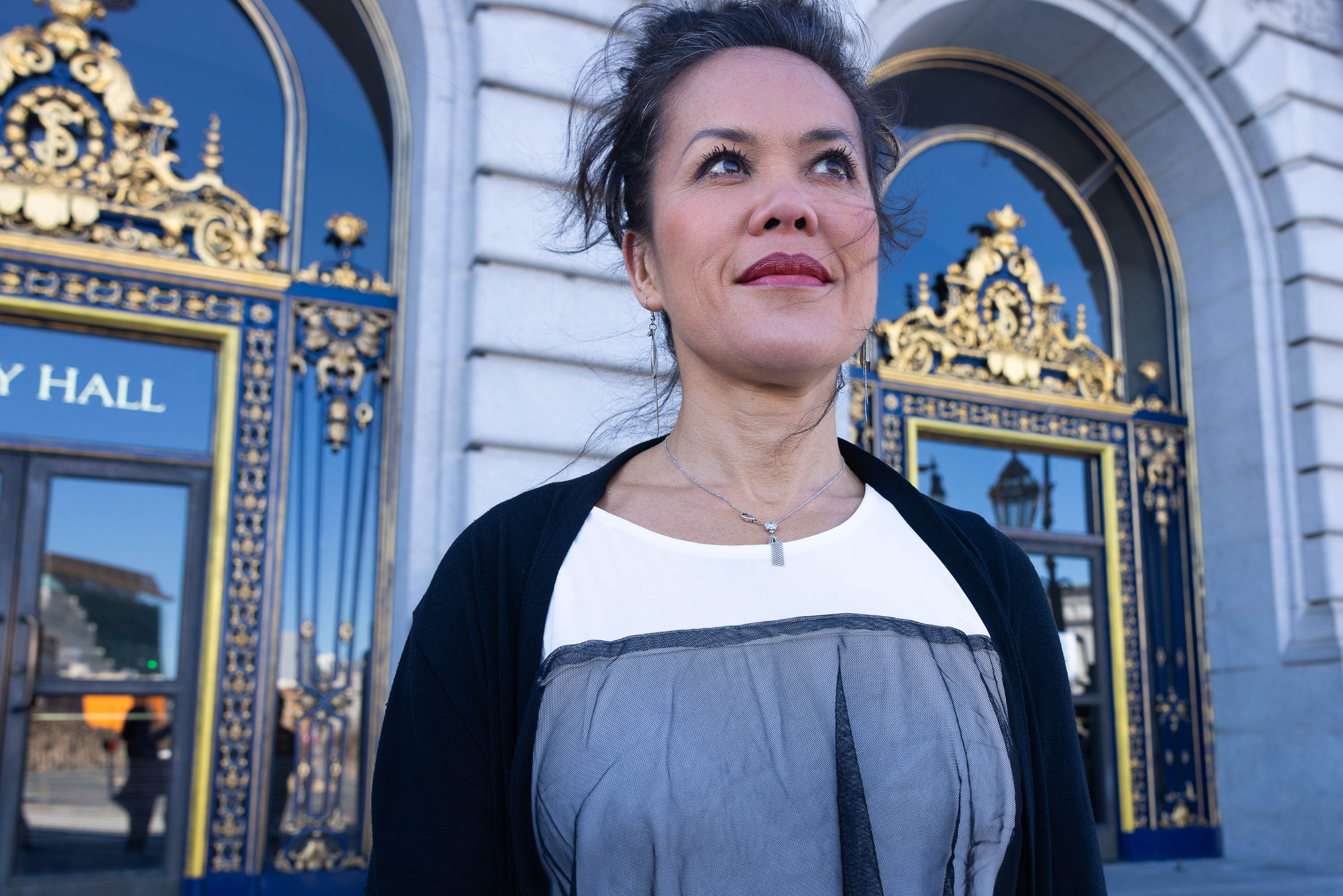 A woman stands before an ornate building with golden detailing, gazing upward thoughtfully.