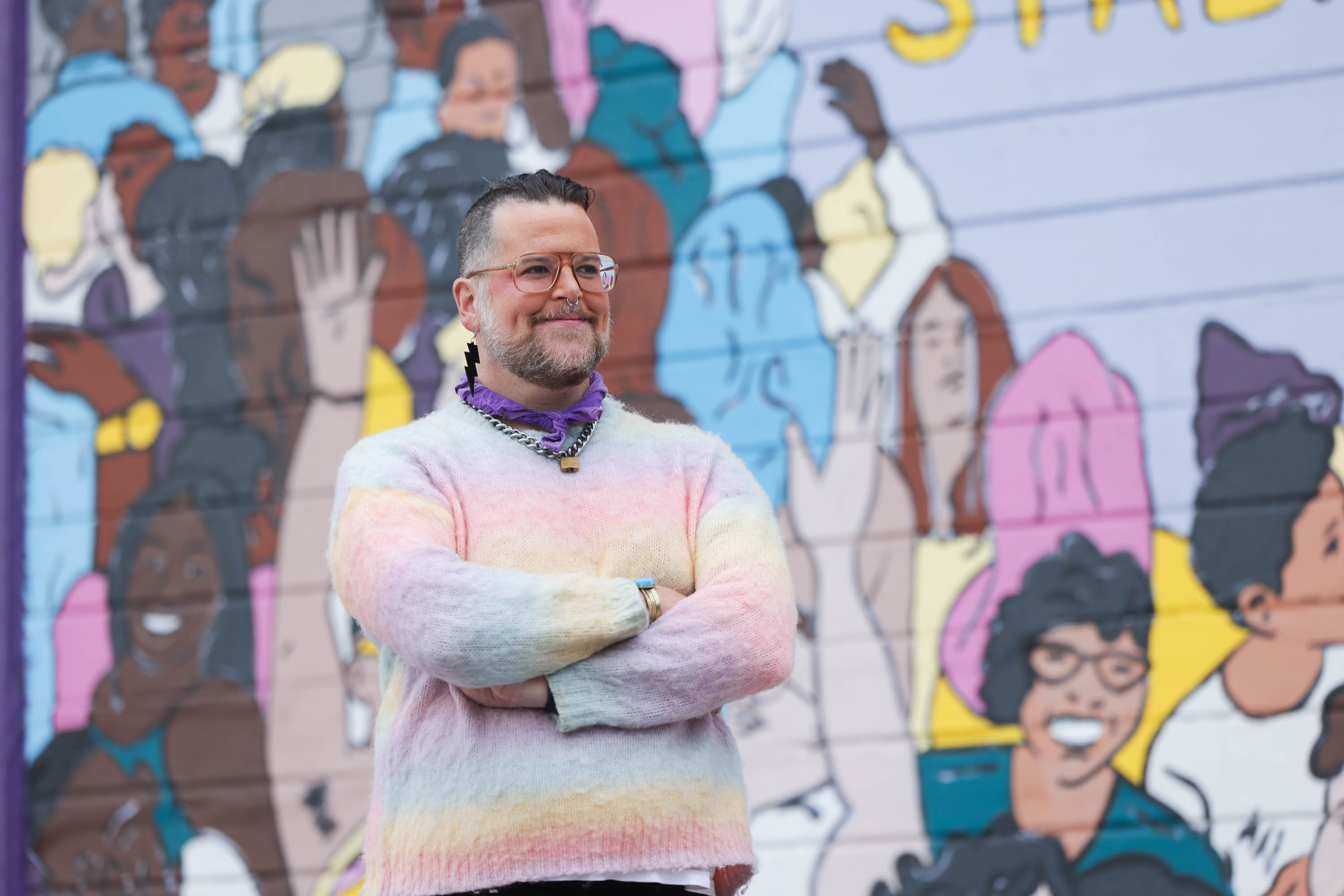 a smiling nonbinary person in a fuzzy pastel sweater crosses their arms in front of a colorful LGBTQ mural. 