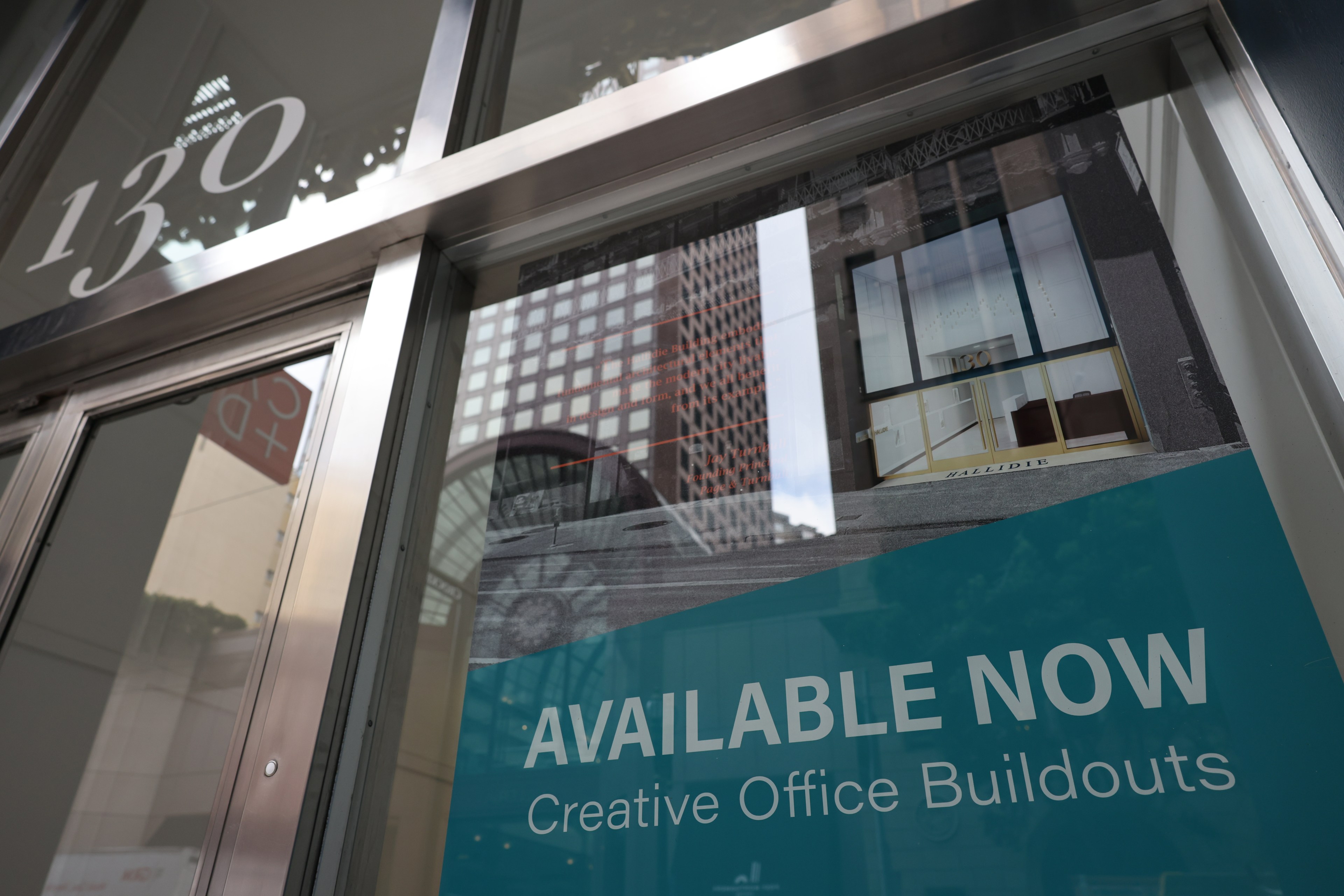 A storefront with &quot;AVAILABLE NOW Creative Office Buildouts&quot; sign, reflecting city buildings.