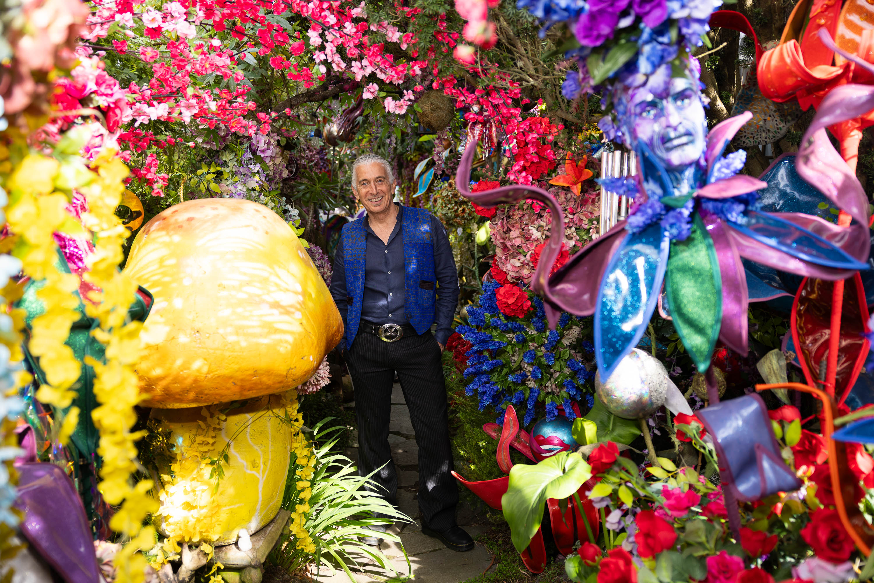 A man stands among colorful flowers and whimsical decorations, including a large fake mushroom.