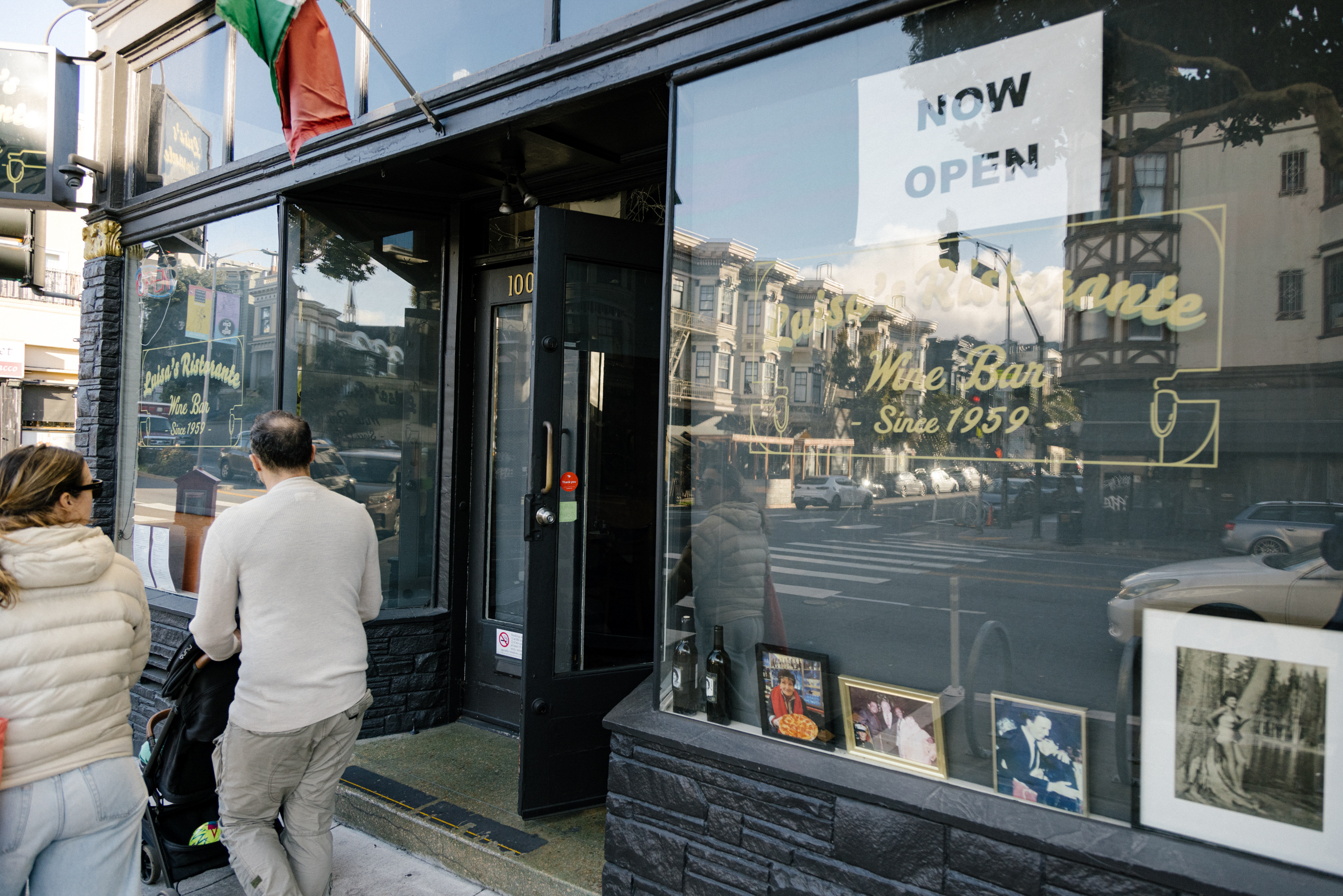 A couple with a stroller stands outside a bar with a &quot;Now Open&quot; sign, as street reflections merge with the interior.