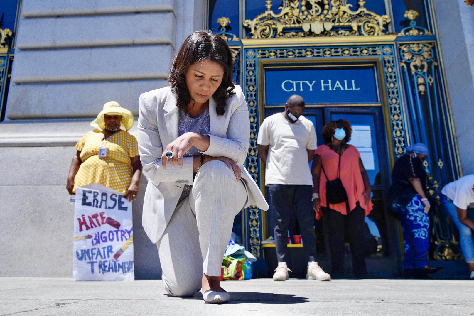 Mayor London Breed kneels on the ground in front of the City Hall entrance, as people hold protest signs in the background.