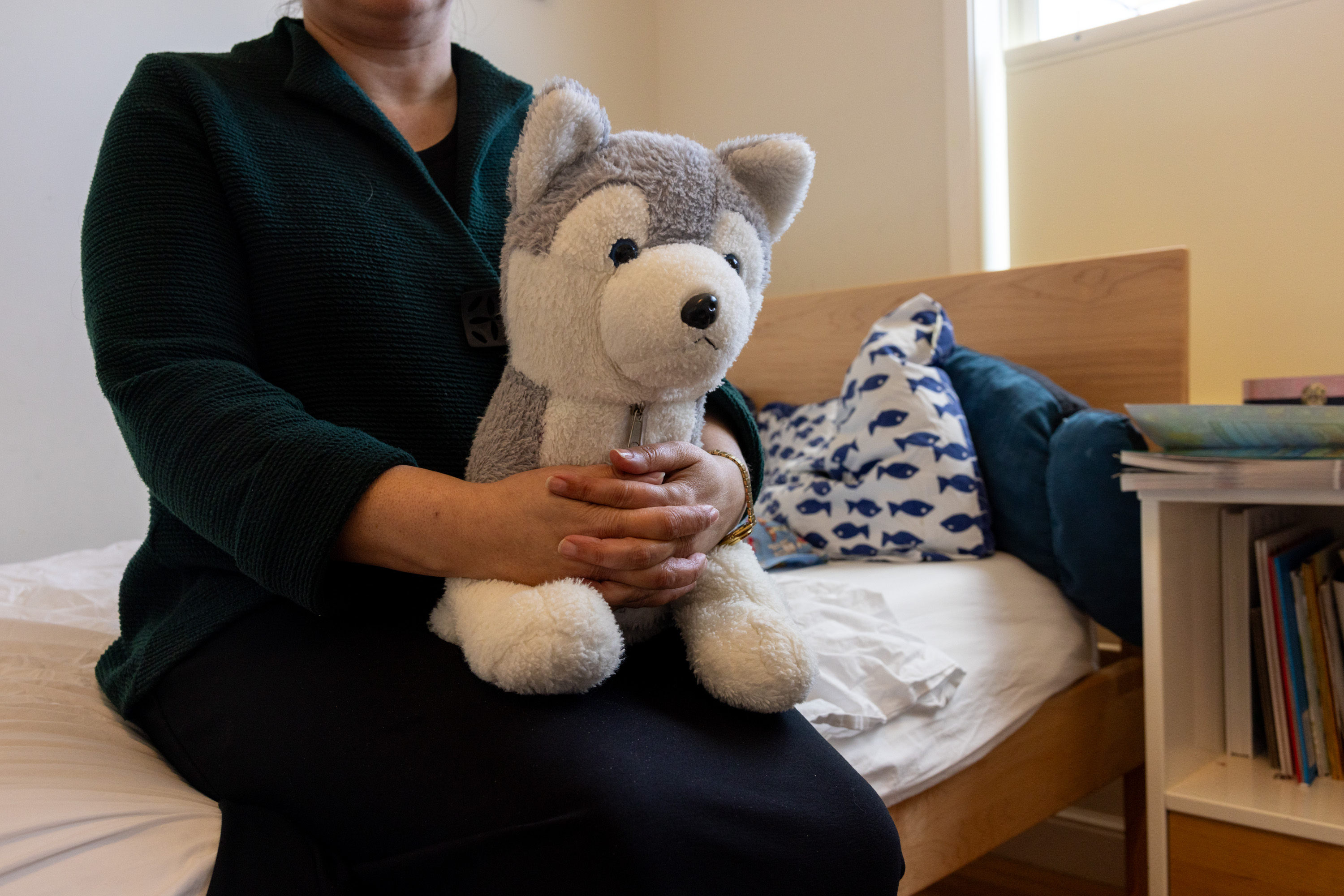 A person is seated holding a plush wolf toy, with a bed and pillow in the background.