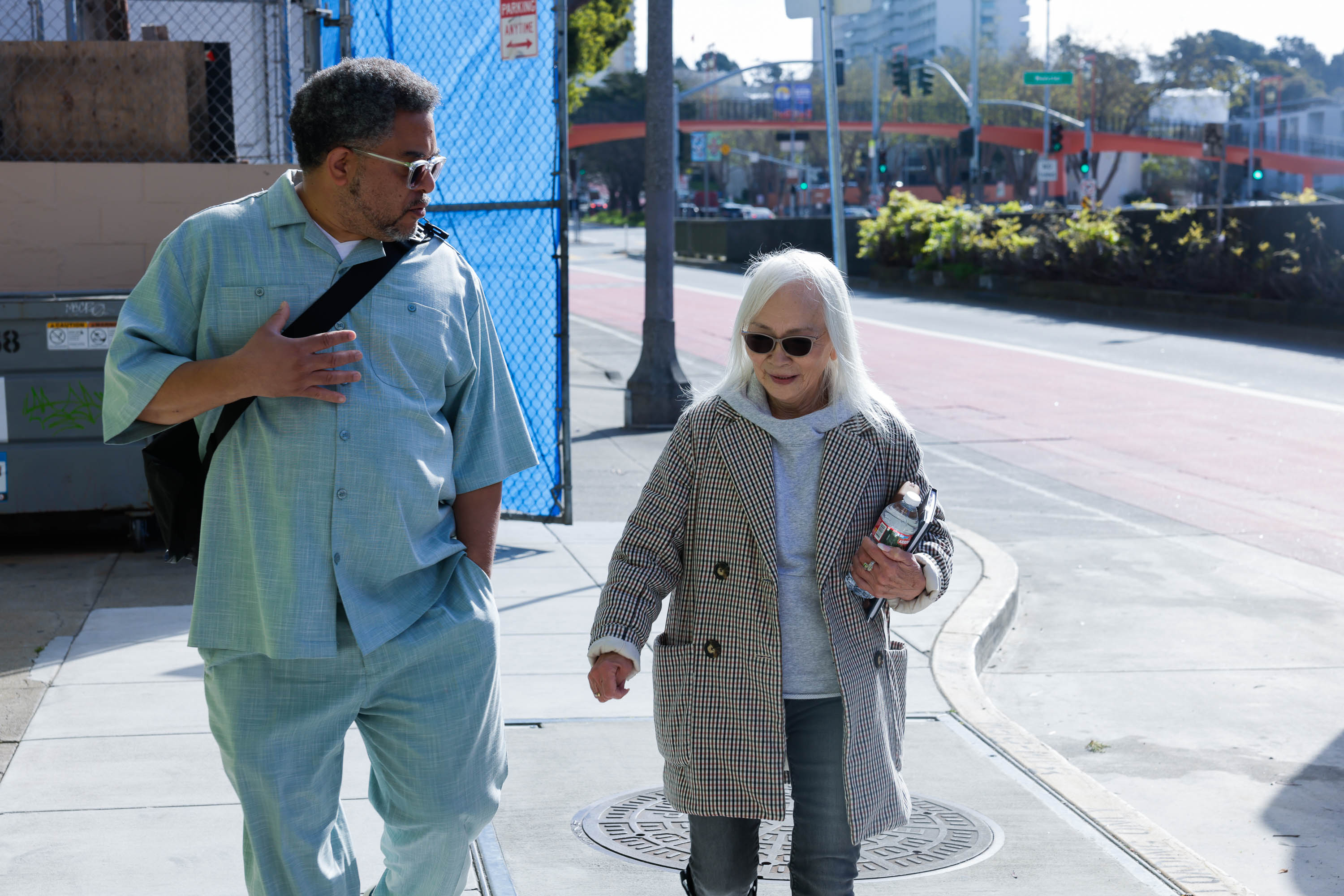 A man and a woman walk on a sunny sidewalk; the man glances aside, the woman carries a bottle.