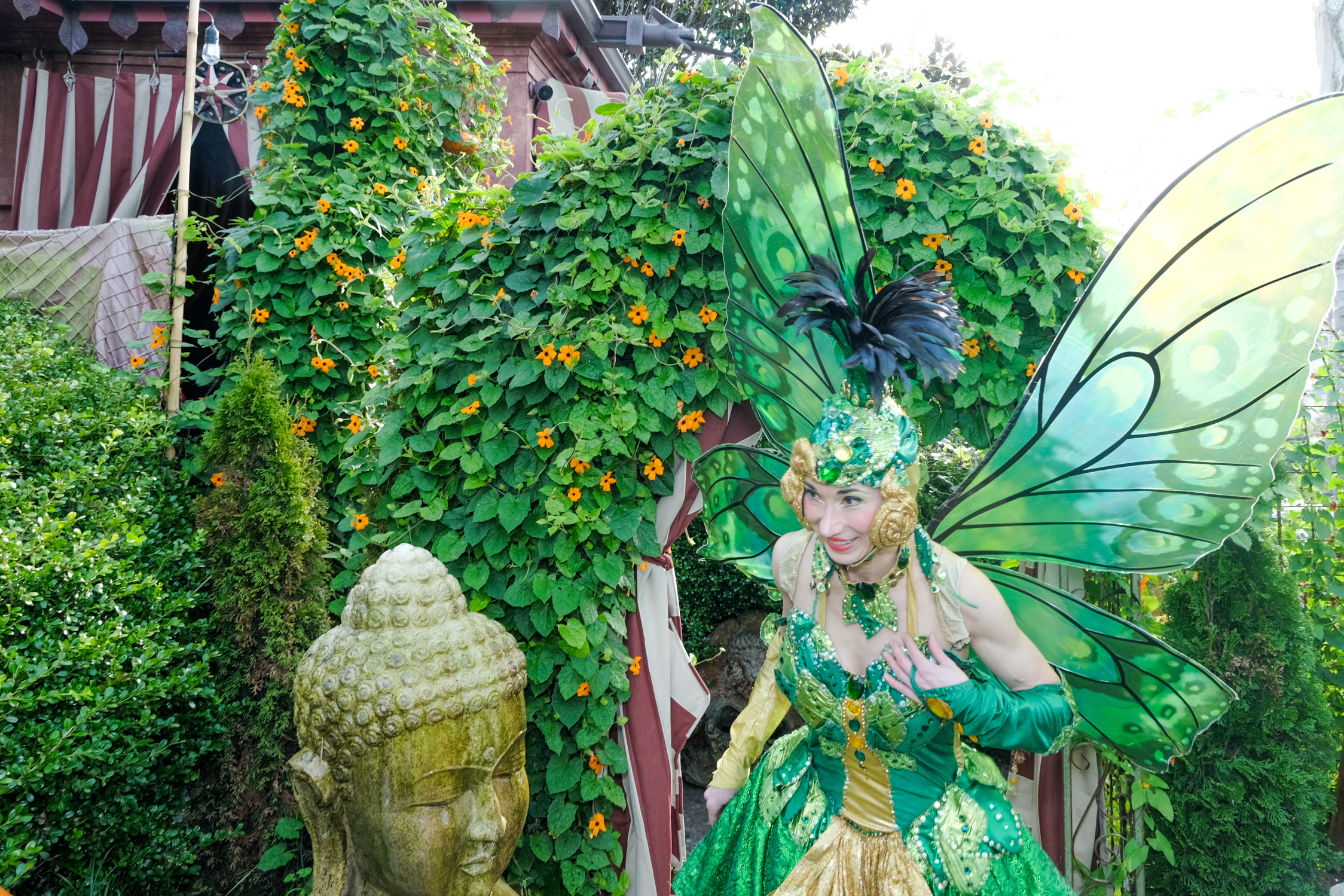 A person dressed as a vibrant green fairy with large wings, amidst lush greenery and orange flowers, next to a stone Buddha head.