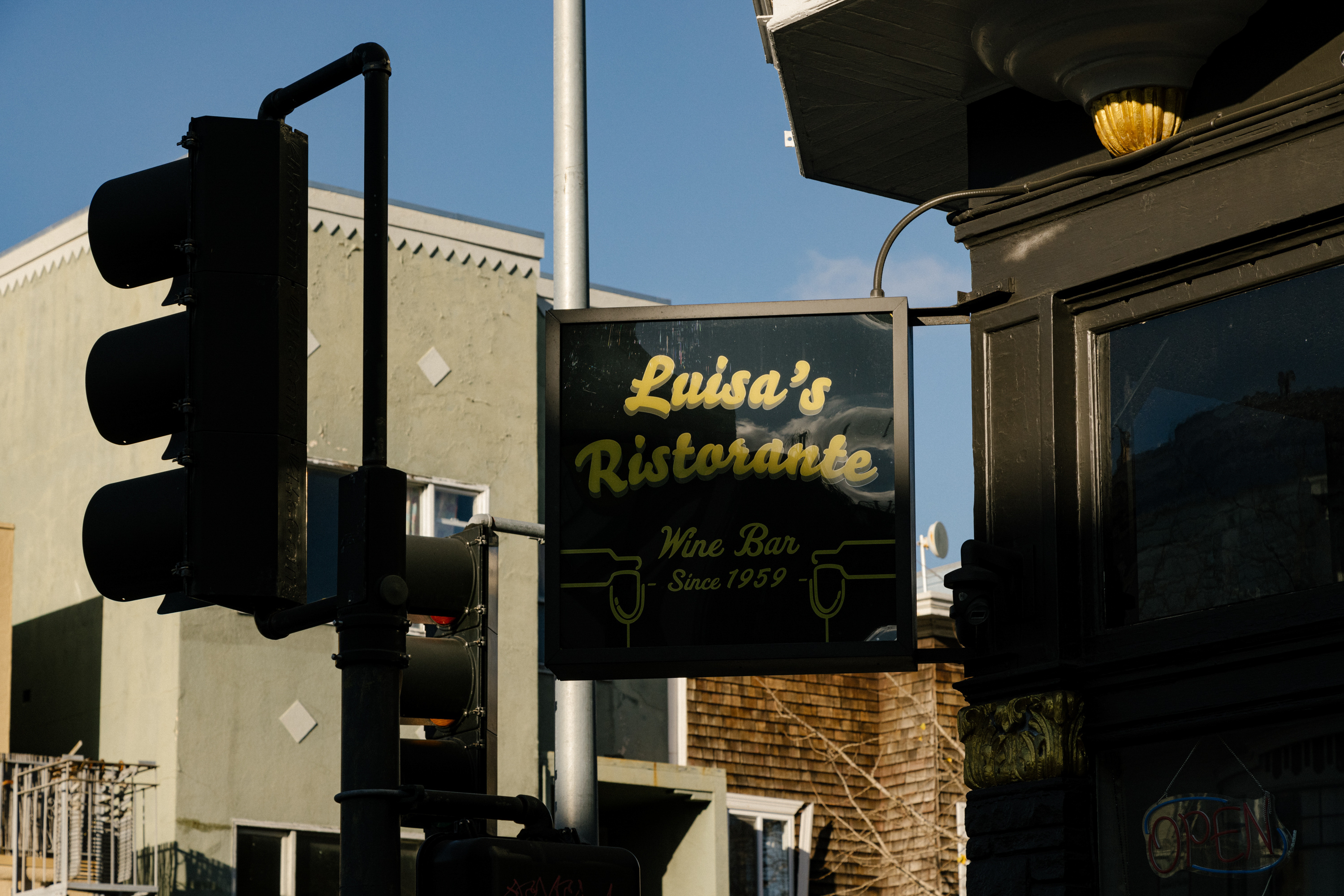 A sign reading &quot;Luisa's Ristorante Wine Bar Since 1959&quot; beside a traffic light, with building and sky in the background.