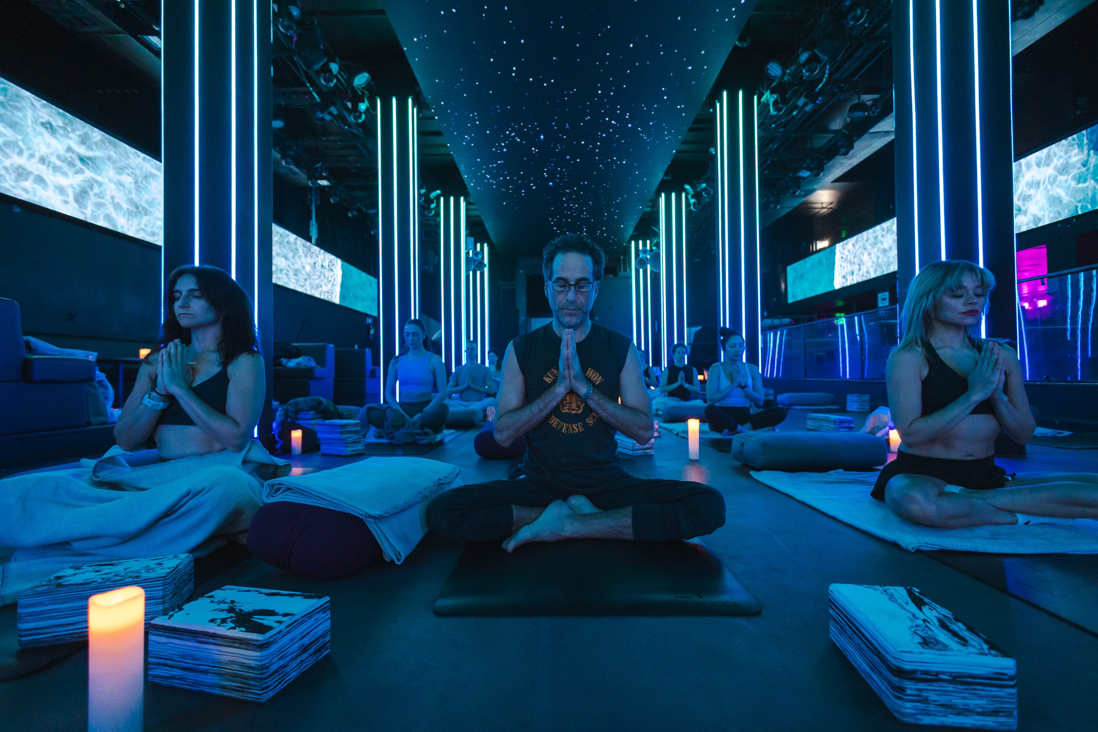 People meditate in a room with blue lighting, LED columns, starry ceiling, and video screens.