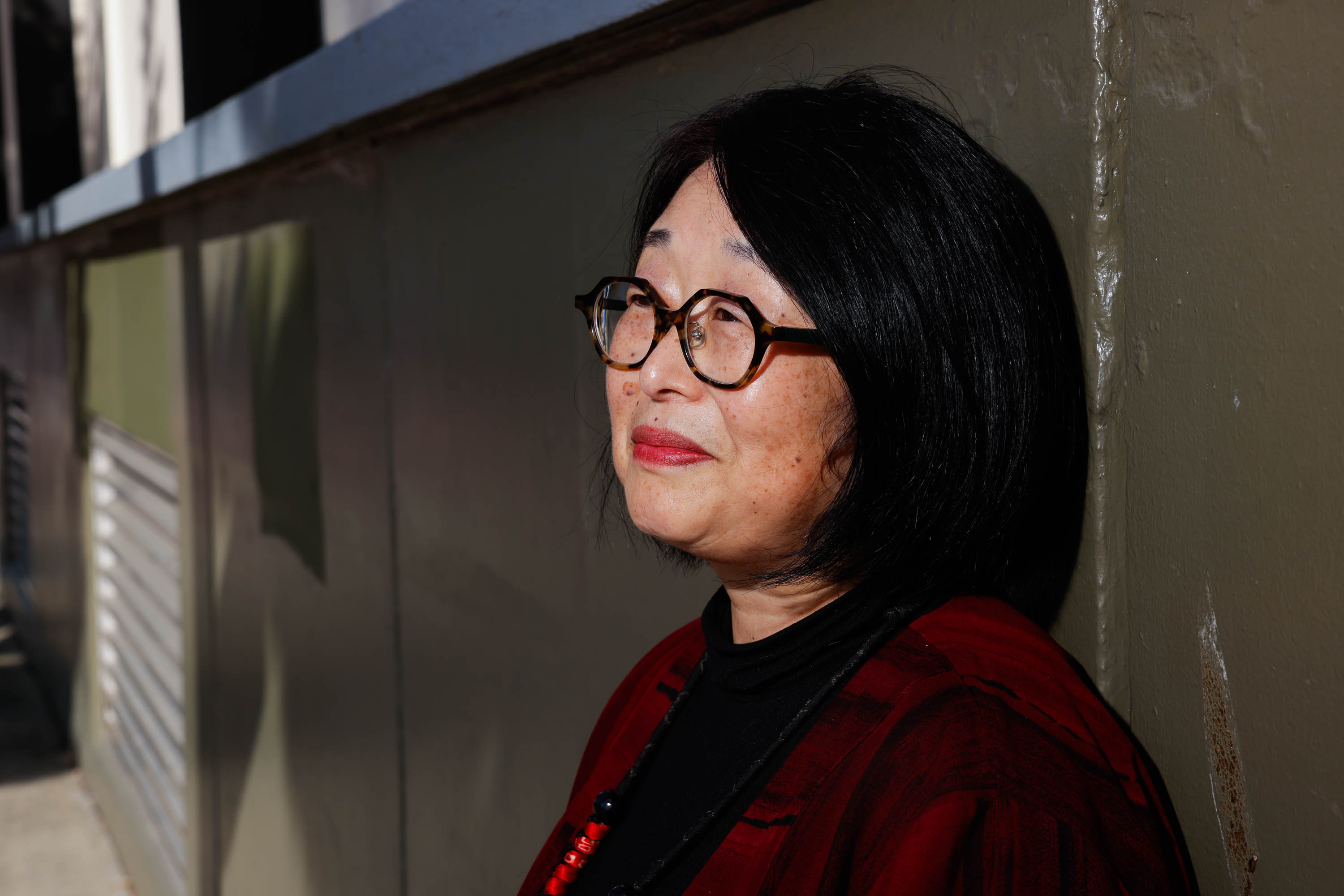 A woman with glasses and black hair gazes away from the camera, against a sunlit wall.