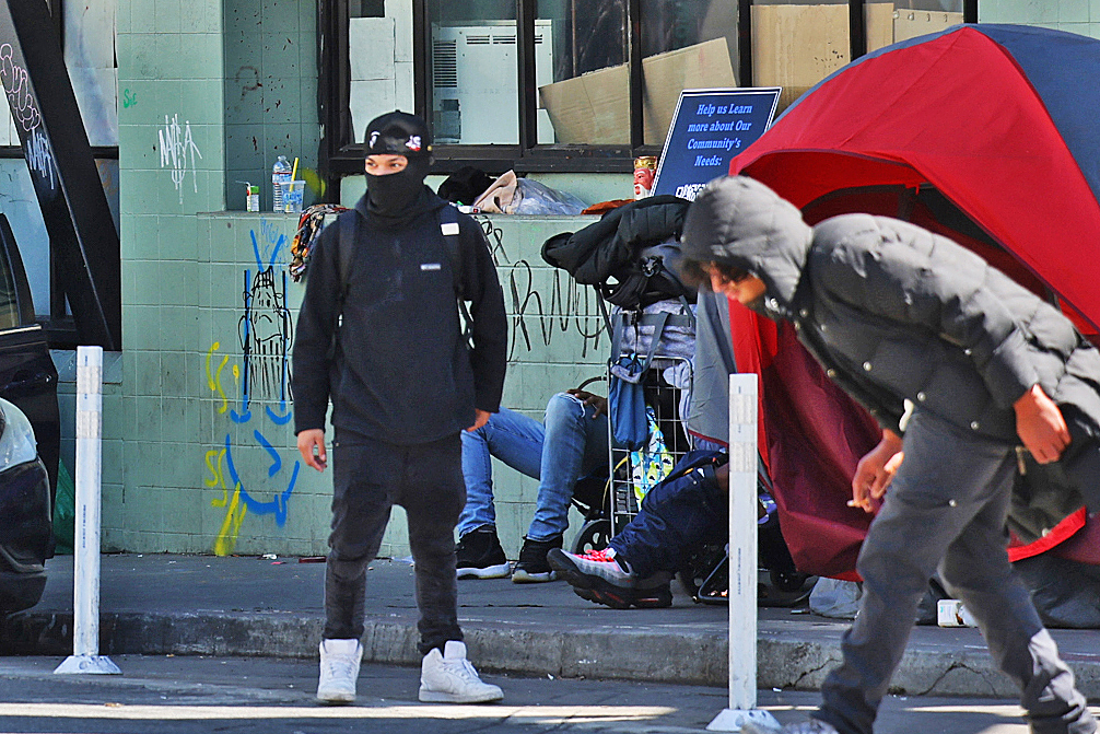 A person in a mask walks by graffiti and a tent on a busy sidewalk.