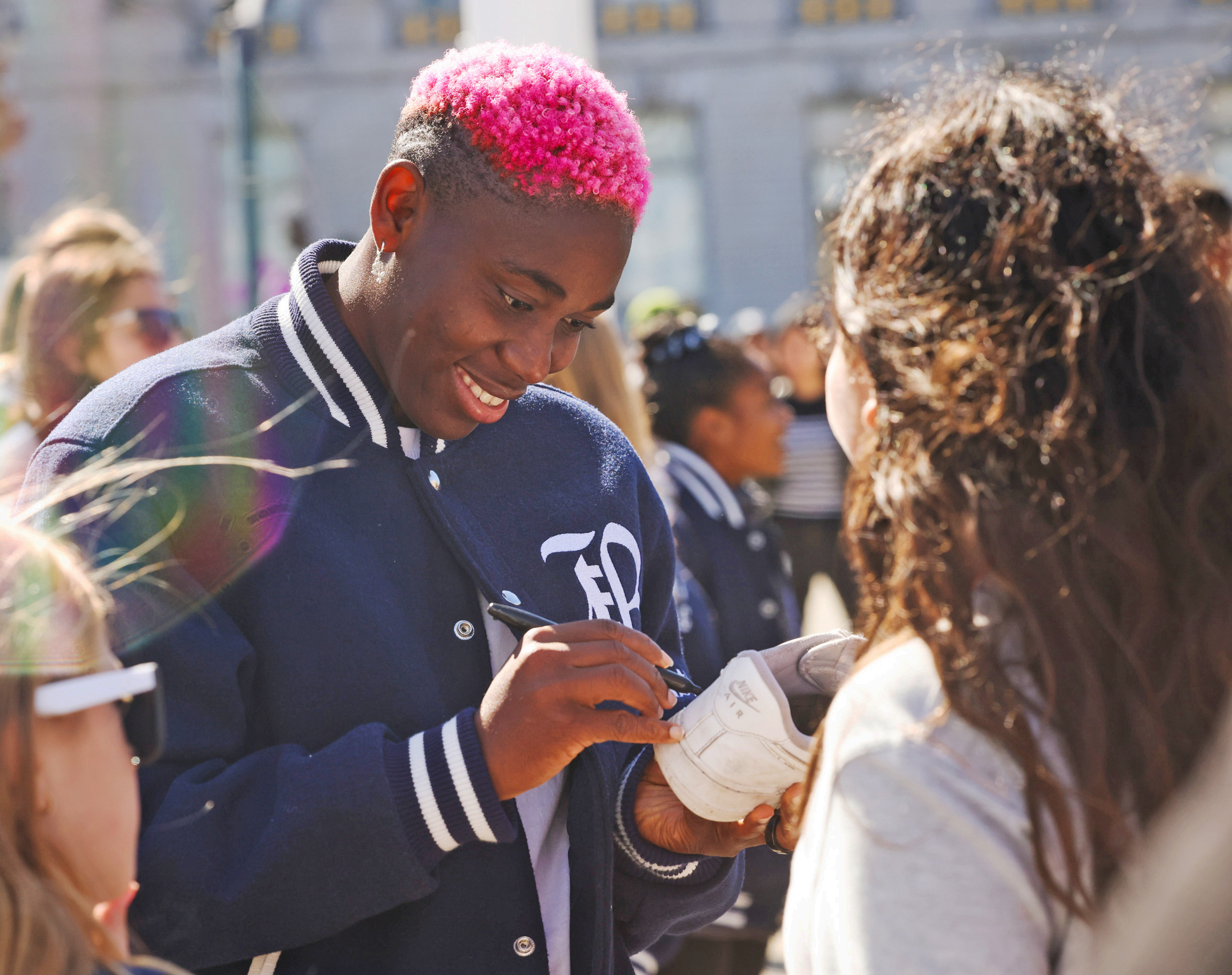 A person with pink hair is smiling and signing a white shoe for a fan outdoors, in a sunny, crowded setting.