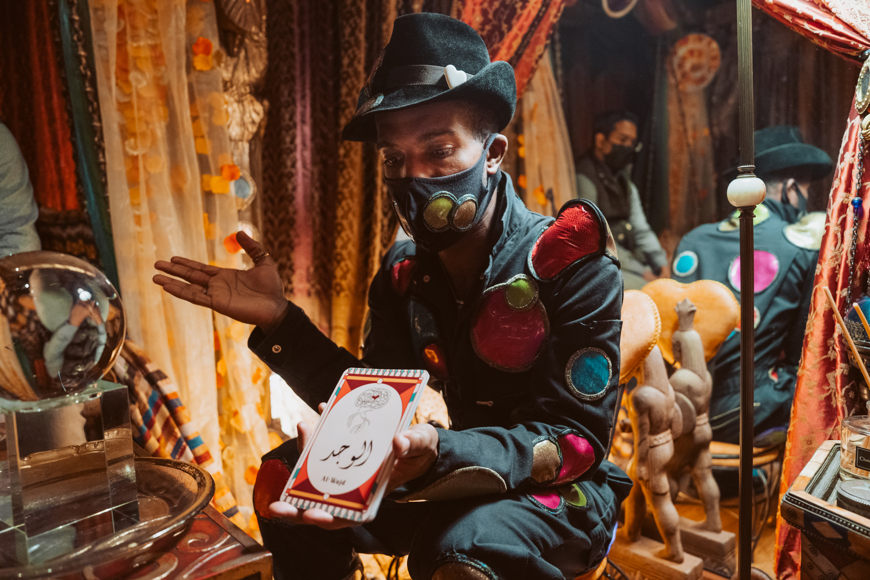 A person in a colorful, patch-adorned jacket showcasing a playing card, wearing a mask and hat, with a reflection and warm tones.