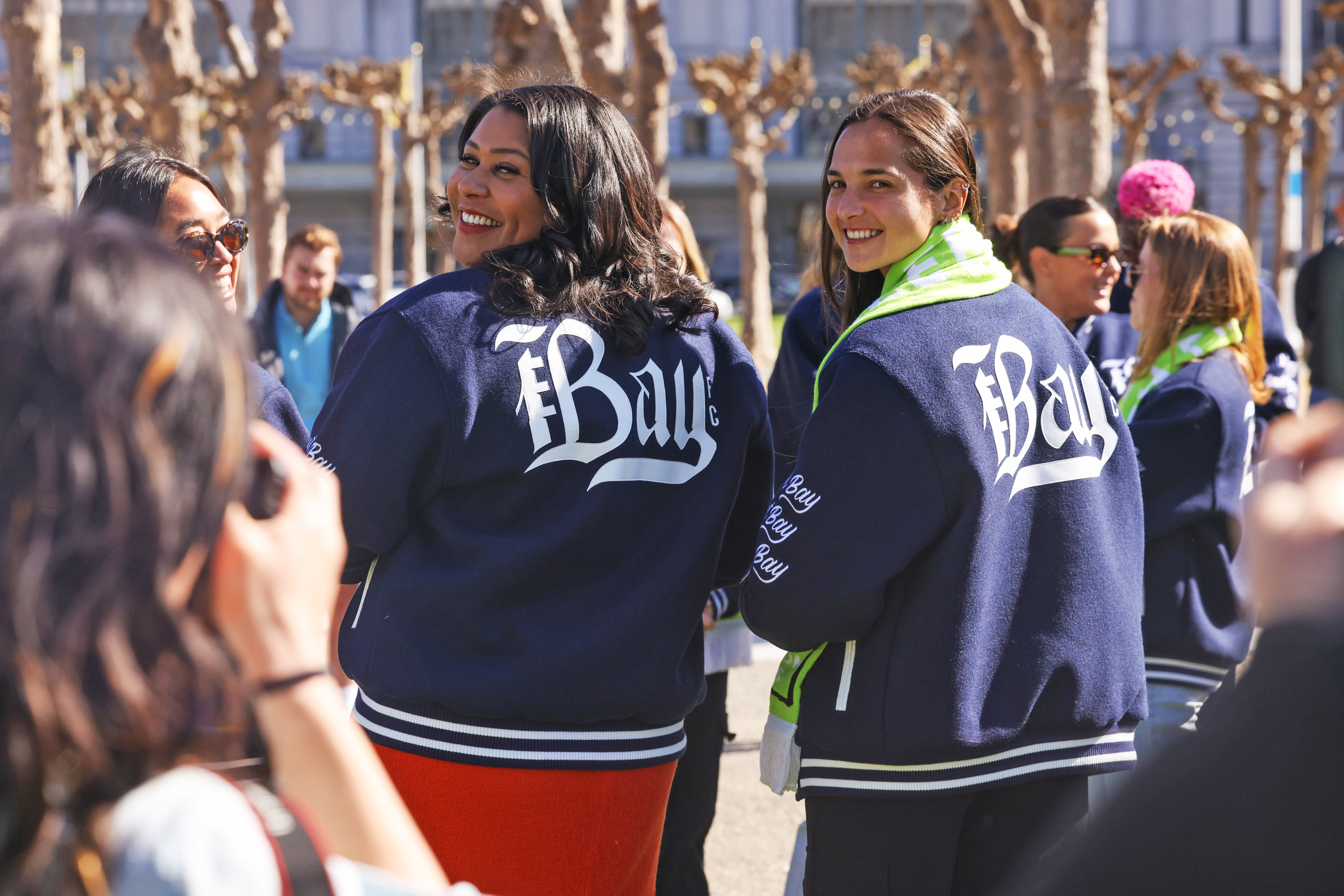 Two smiling women in &quot;#Bay&quot; jackets amidst a sunny outdoor gathering.