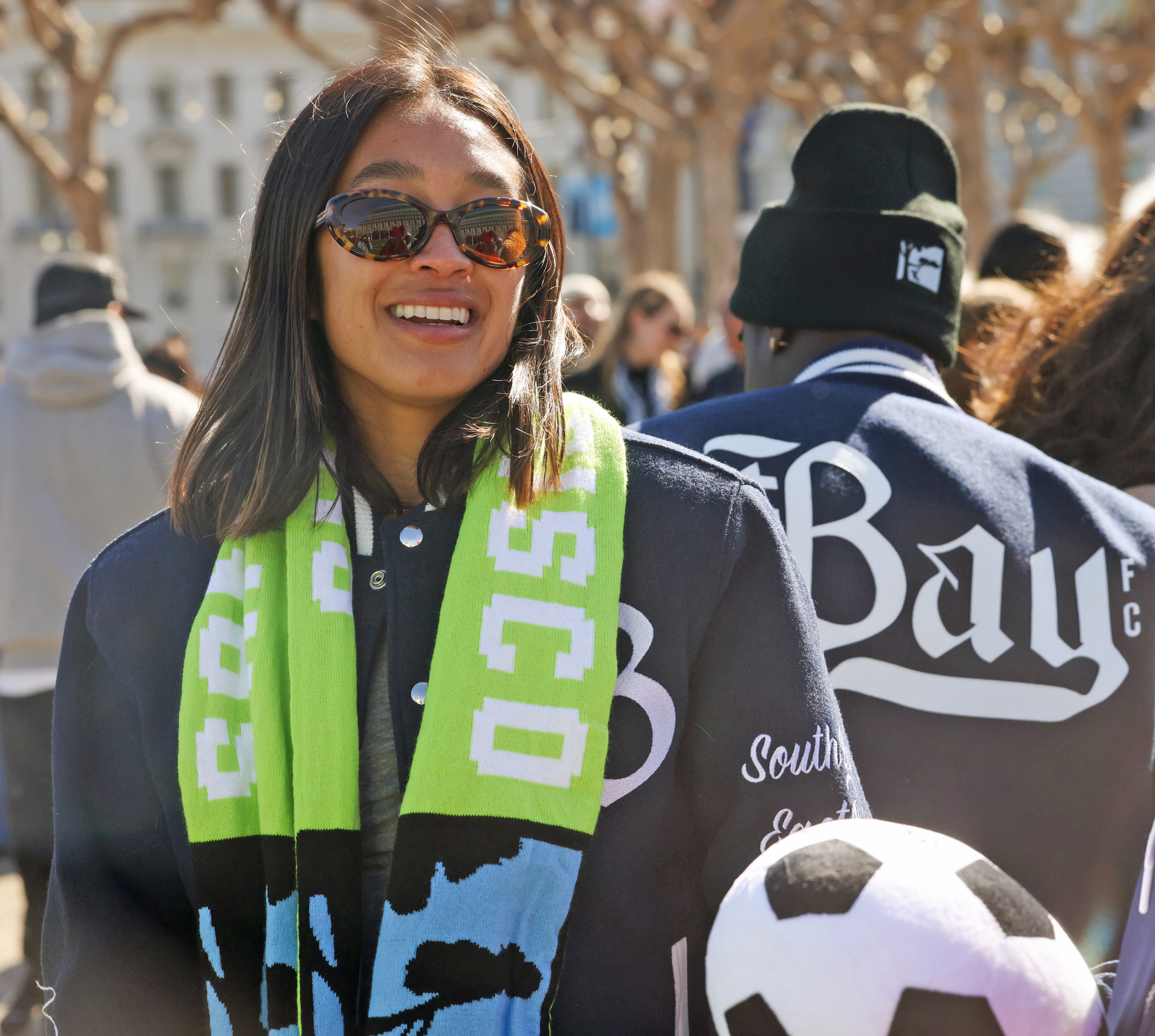 A smiling woman with sunglasses, a colorful scarf, and a soccer ball, with people wearing branded attire in the background.