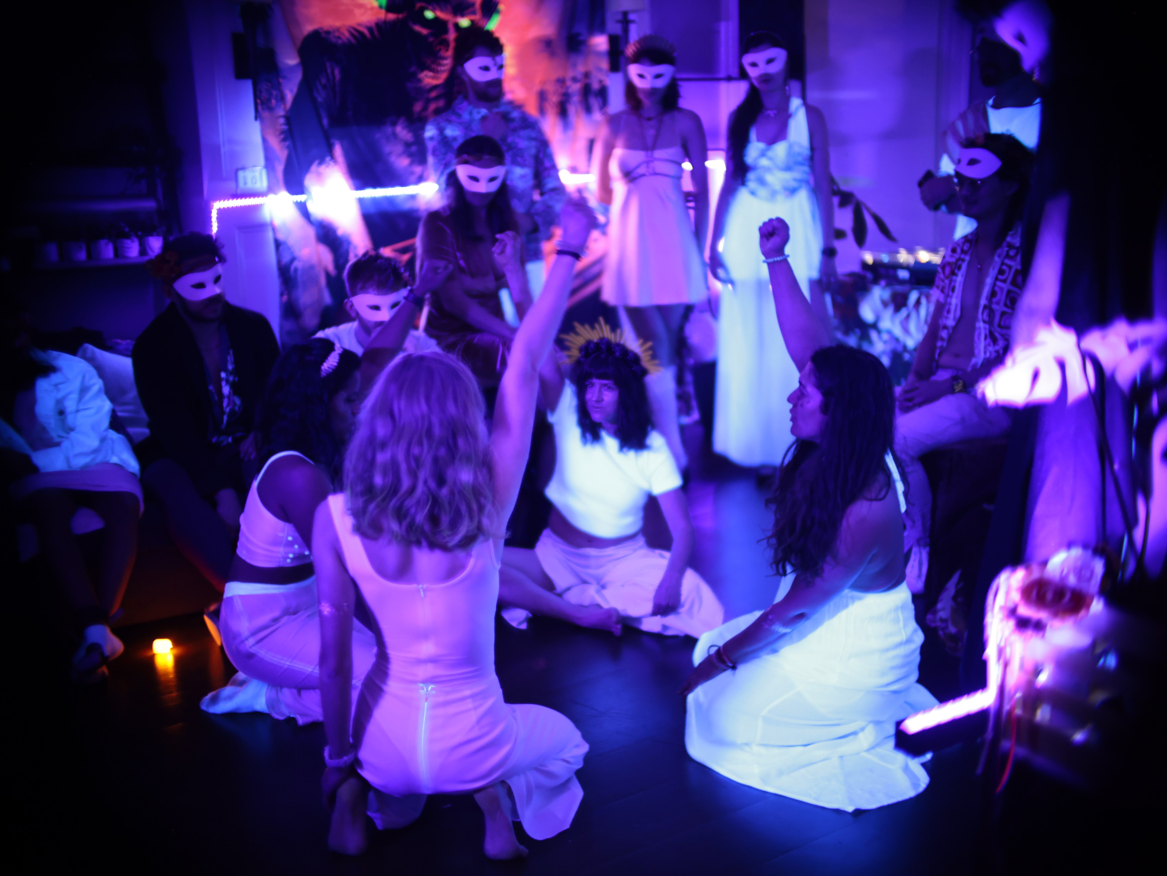 People in a dimly lit room with masks sit in a circle with two women in the center, creating a mysterious vibe.