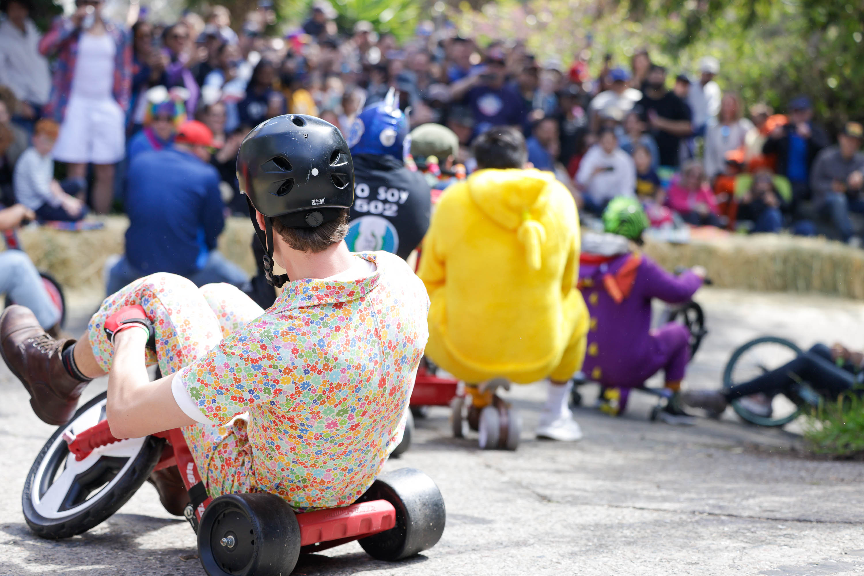 People in costumes race on toy tricycles before a watching crowd.