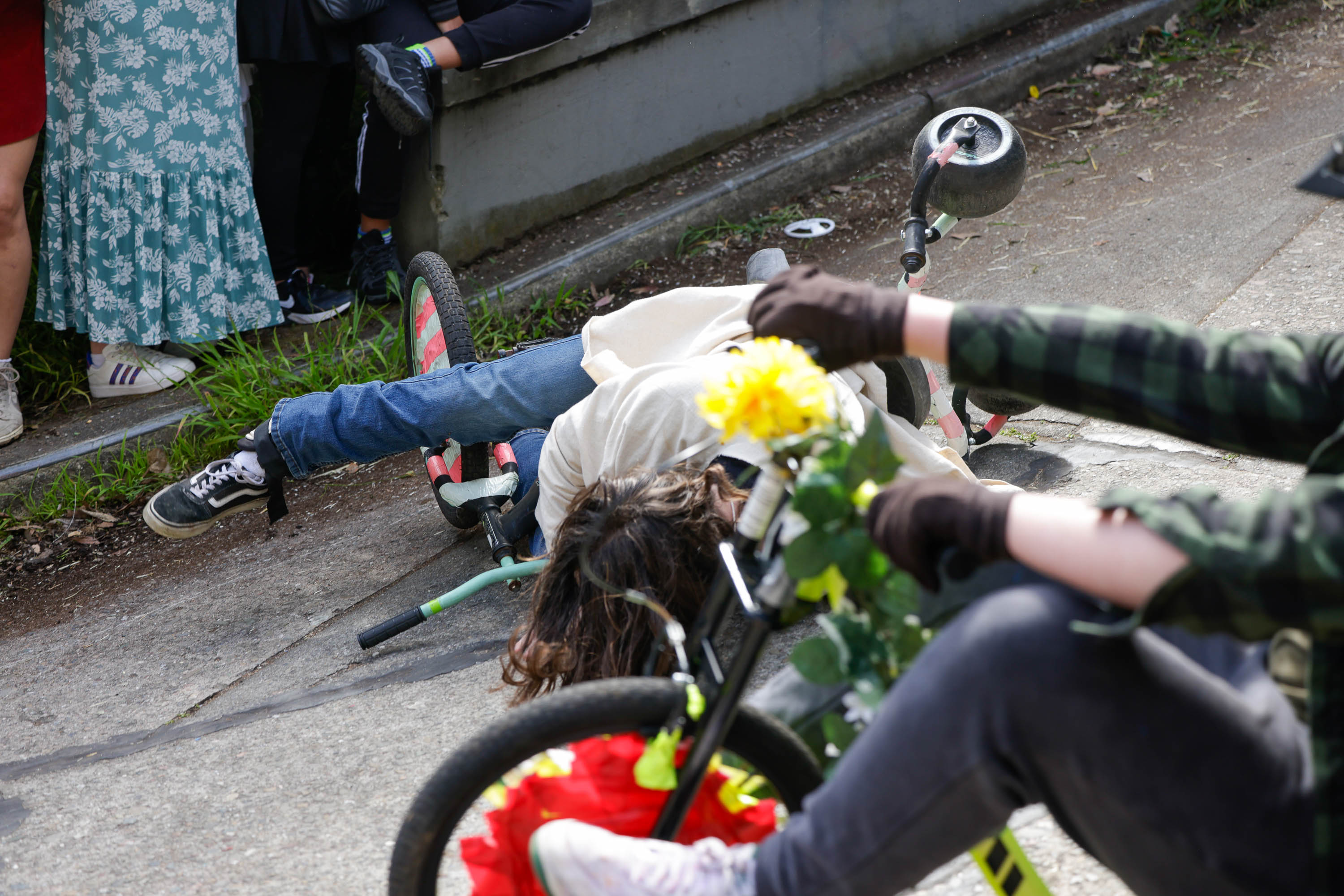 A person lies on the ground beside an overturned bike, with onlookers and flowers visible.