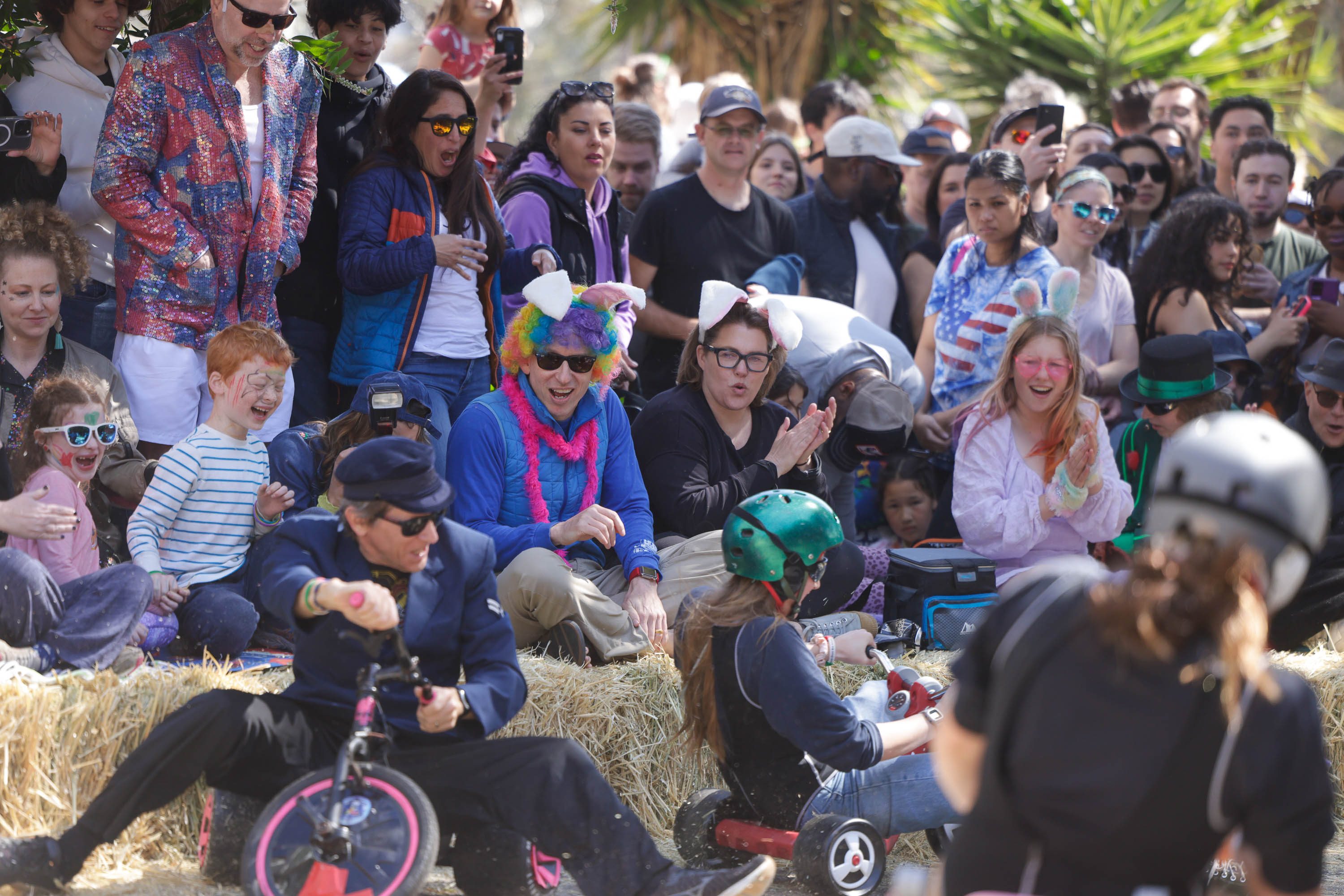 A crowd watches people on tiny bikes; some wear colorful, quirky outfits and helmets.