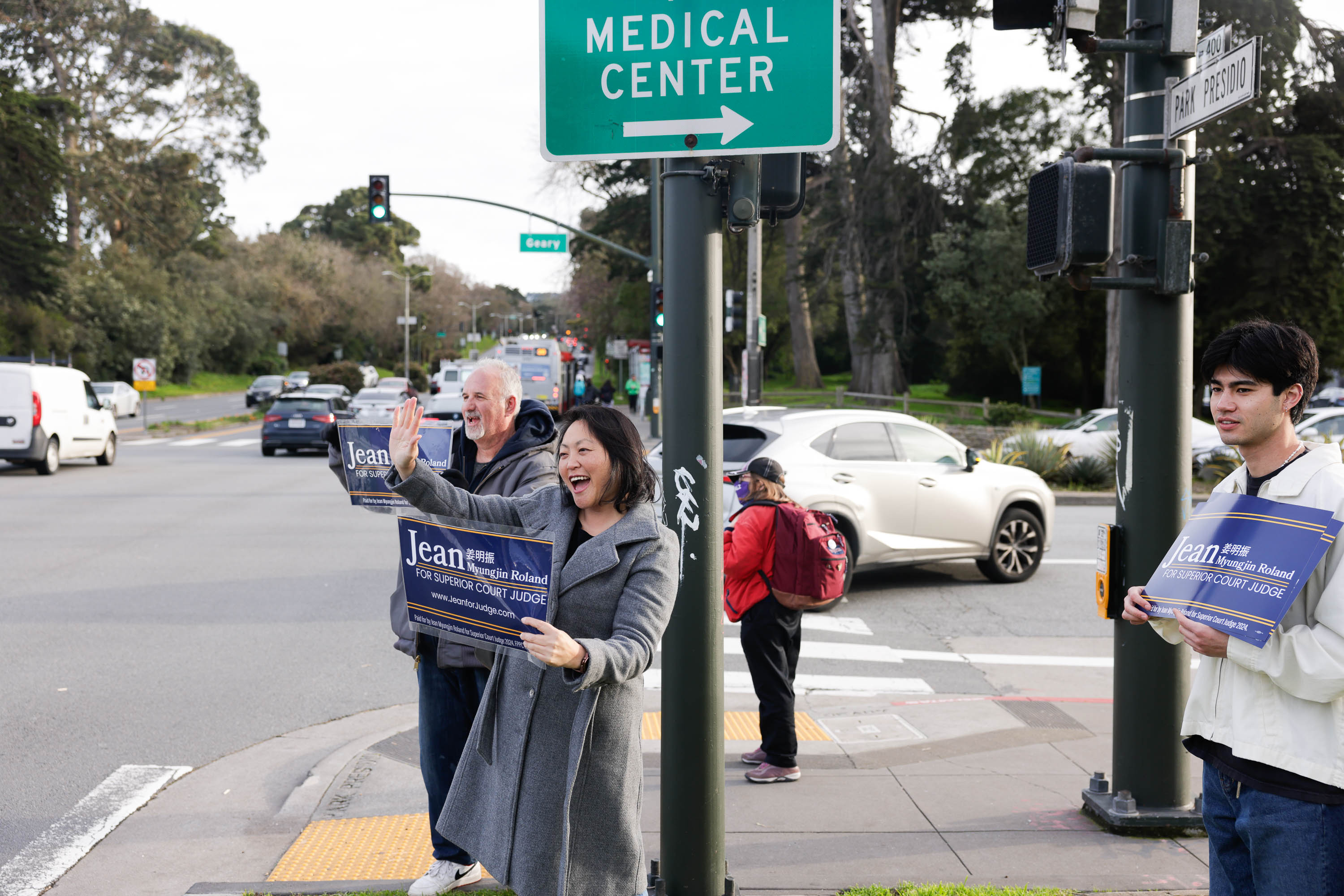 Three people campaign at a street corner near a &quot;Medical Center&quot; sign, holding election signs for a judicial candidate.