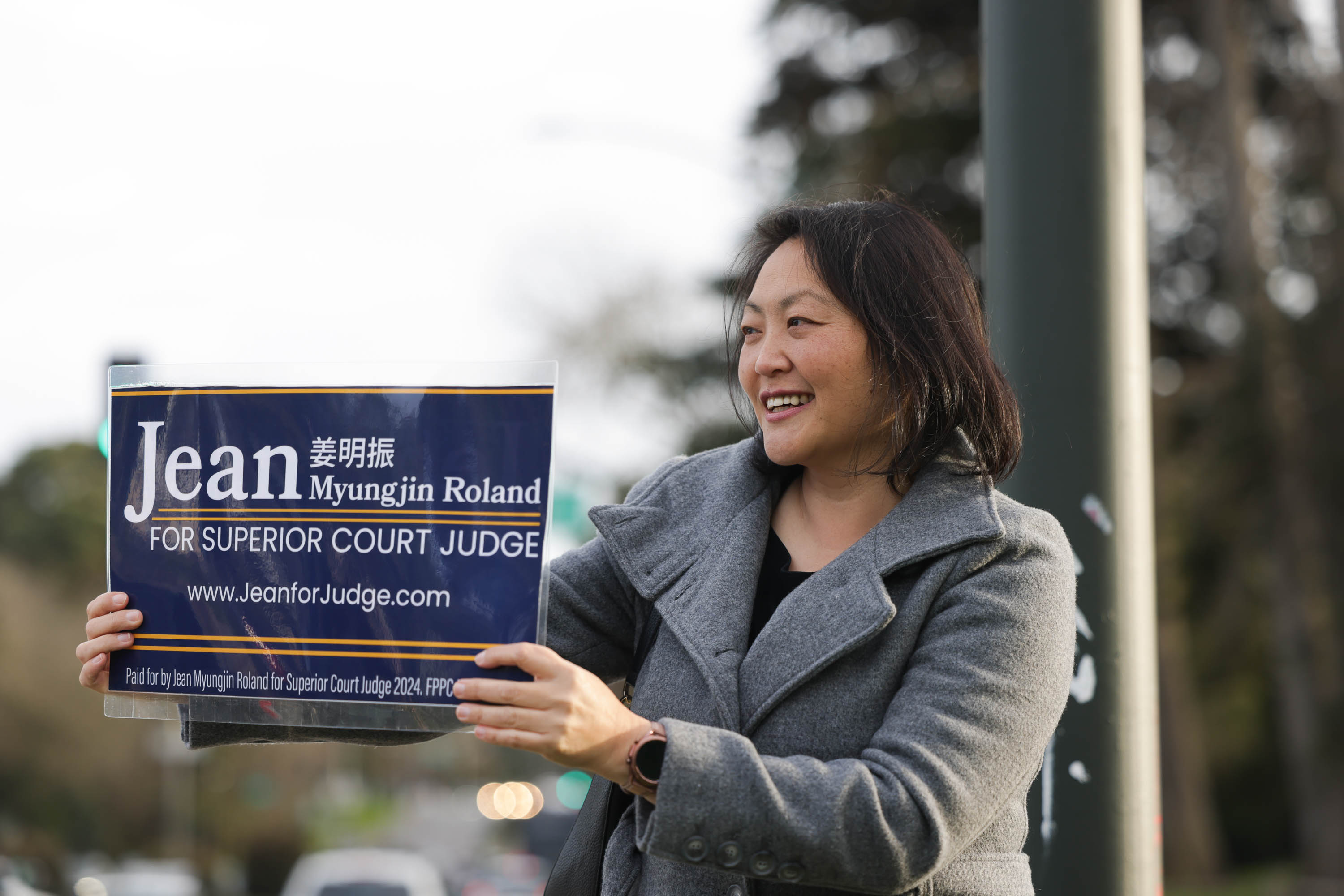 A smiling woman holds a campaign sign for &quot;Jean Myungjin Roland for Superior Court Judge.&quot;