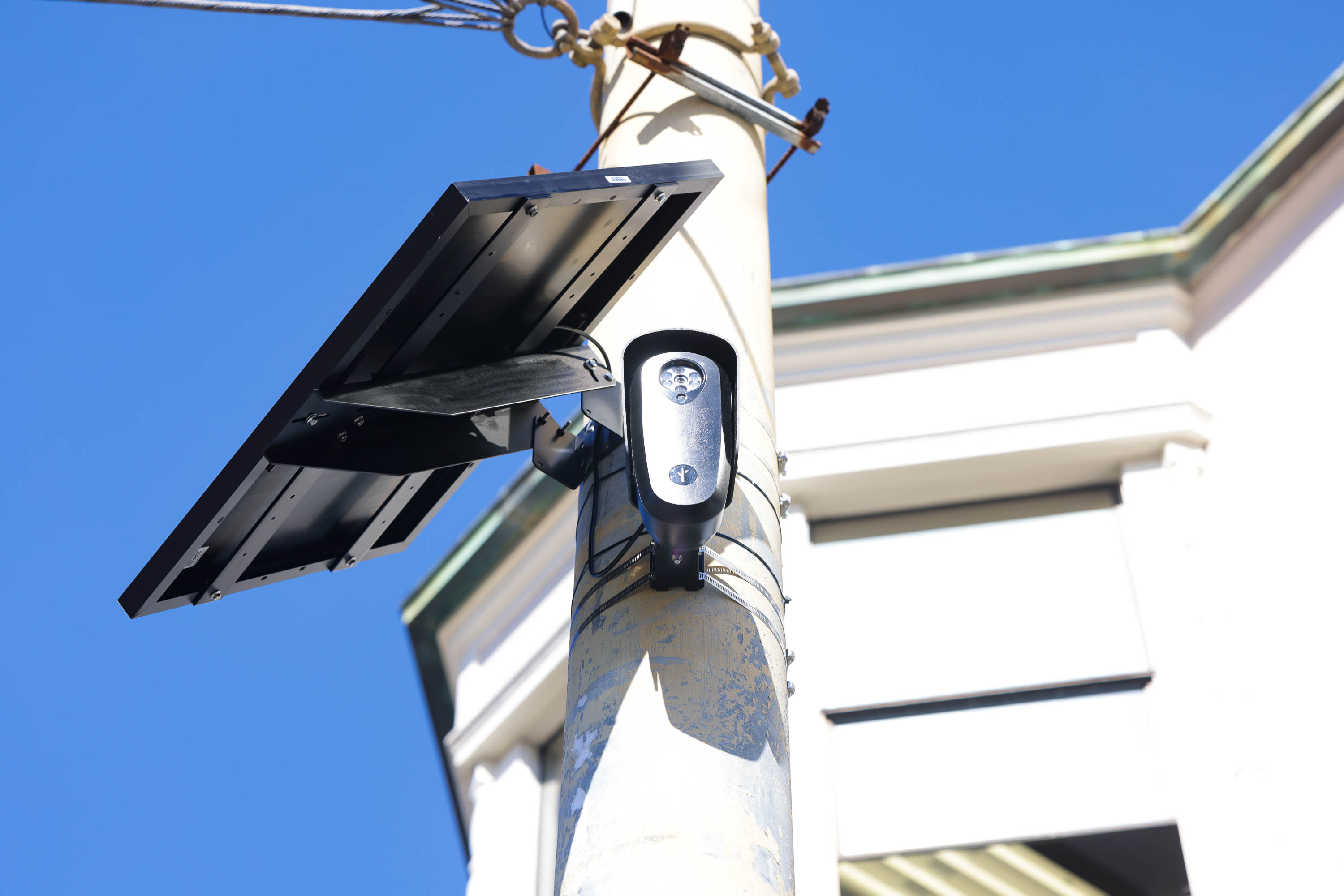 A surveillance camera is mounted on a tall pole next to a solar panel. The sky is clear and blue, with a building partially visible in the background.