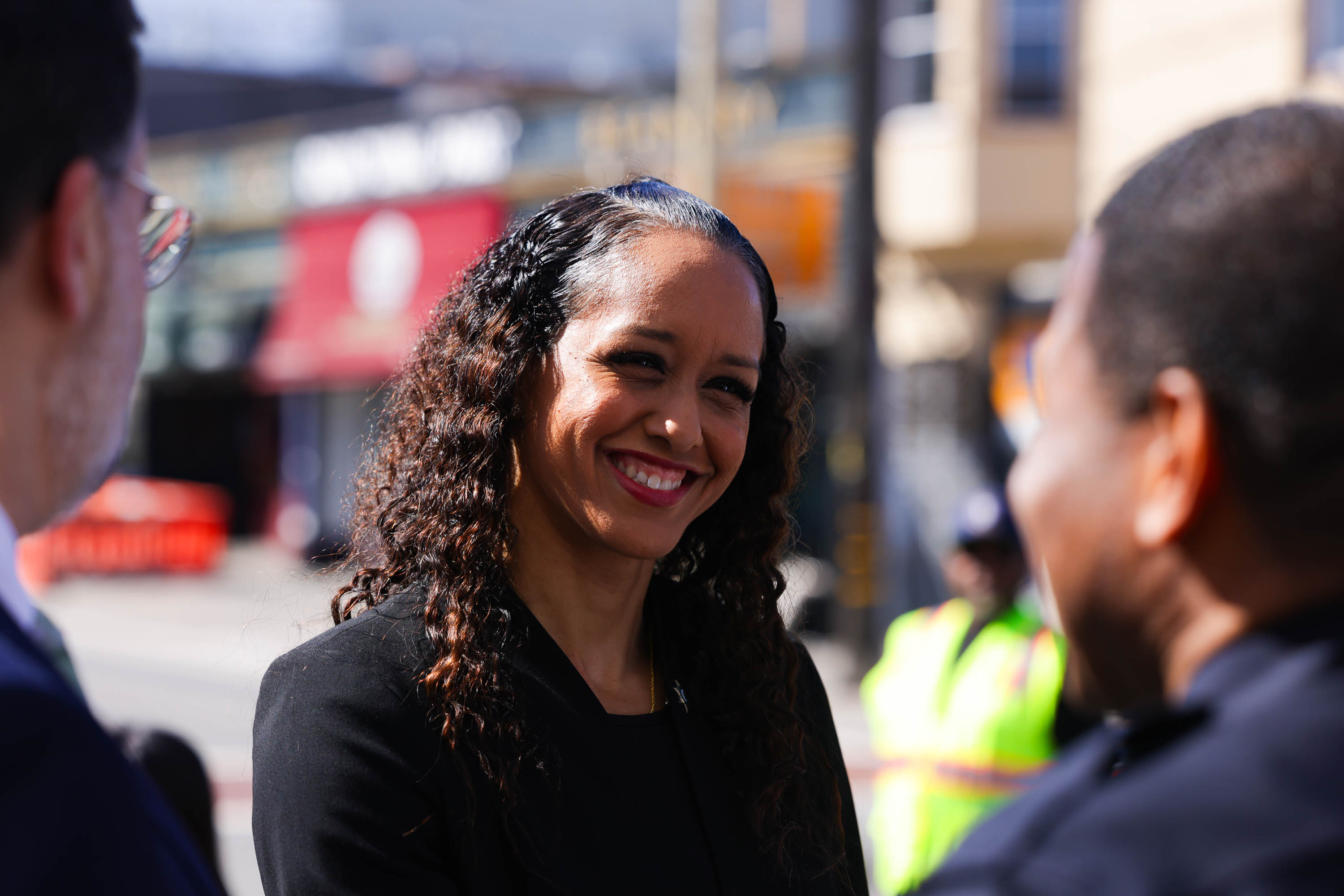 A woman with curly hair smiles while interacting with two people on a sunny day in an urban setting with a red storefront in the background.