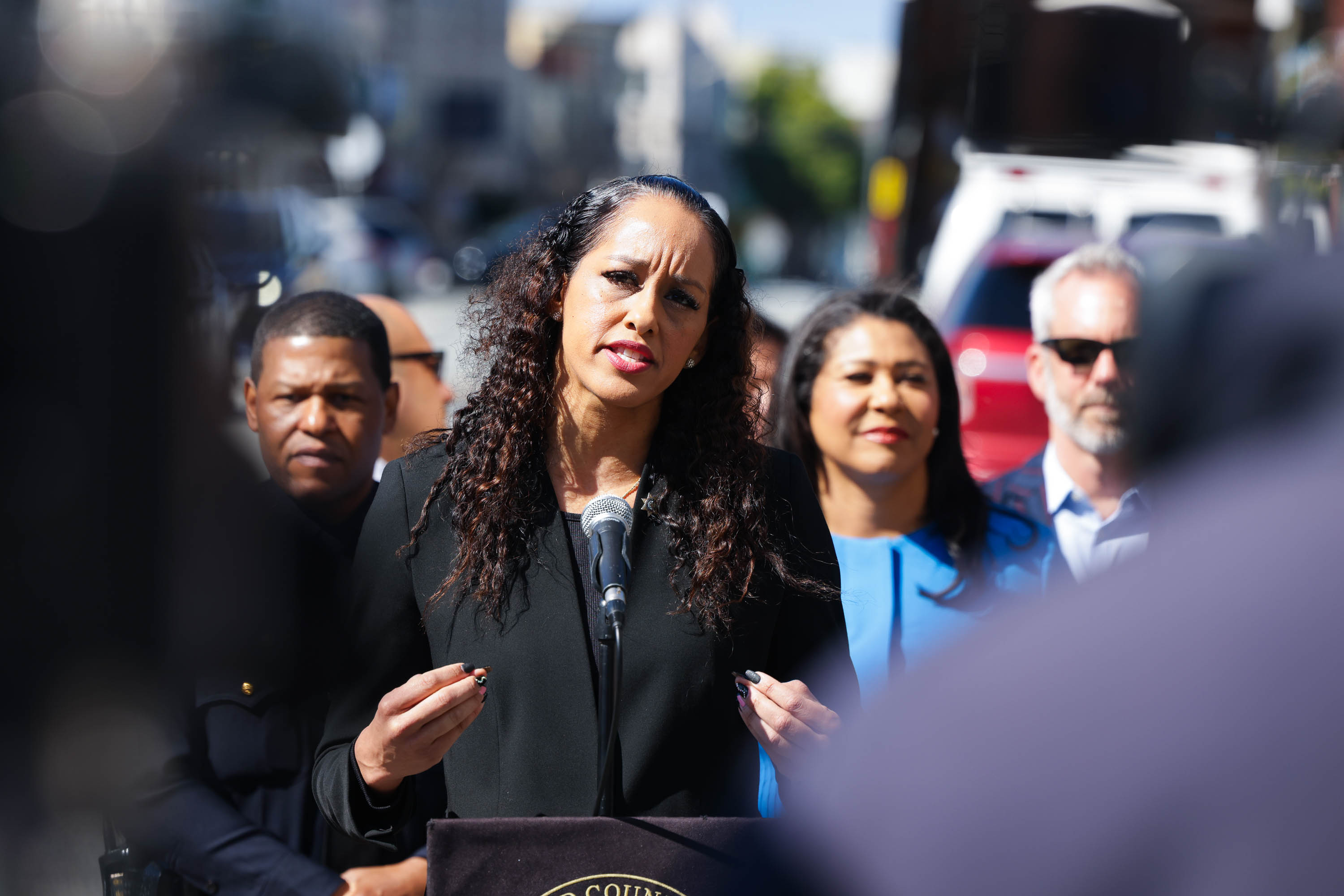 A woman with curly hair speaks passionately at a microphone outdoors, surrounded by people including a man in sunglasses and a woman in a blue outfit.