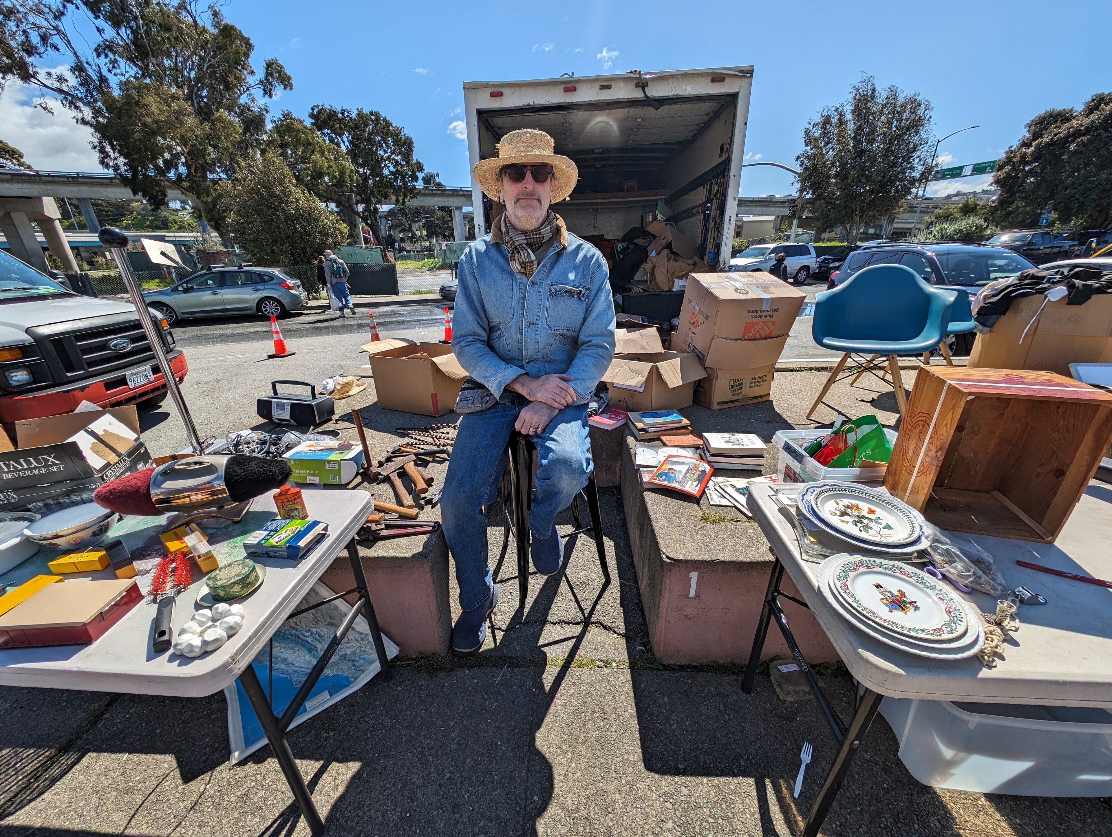 A man wearing sunglasses, a straw hat and denim jacket and jeans sits on a stool surrounded by vintage goods for sale.