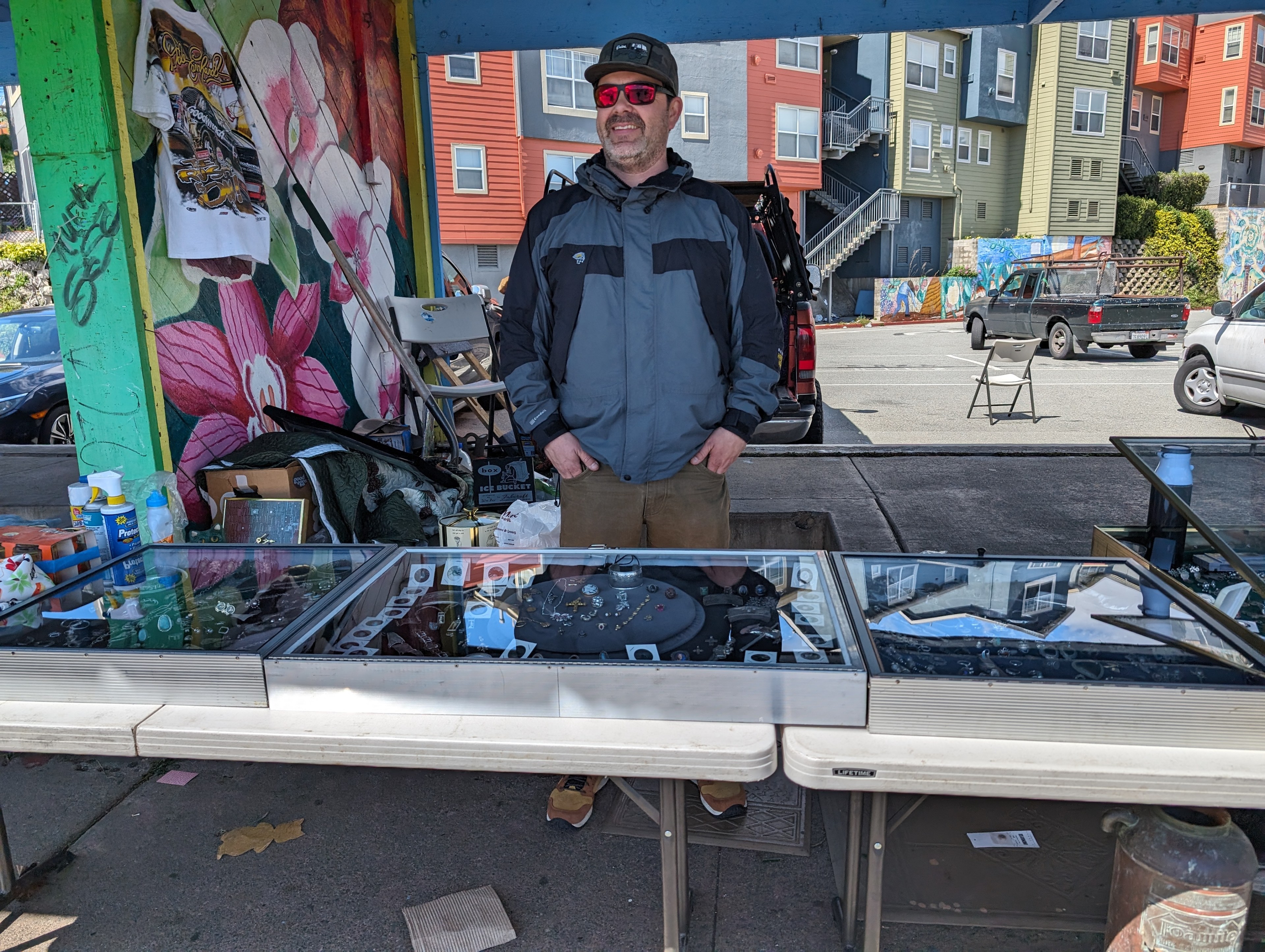 A man in a gray and black jacket, black cap and sunglasses stands behind a table laden with metal cases containing jewelry for sale.